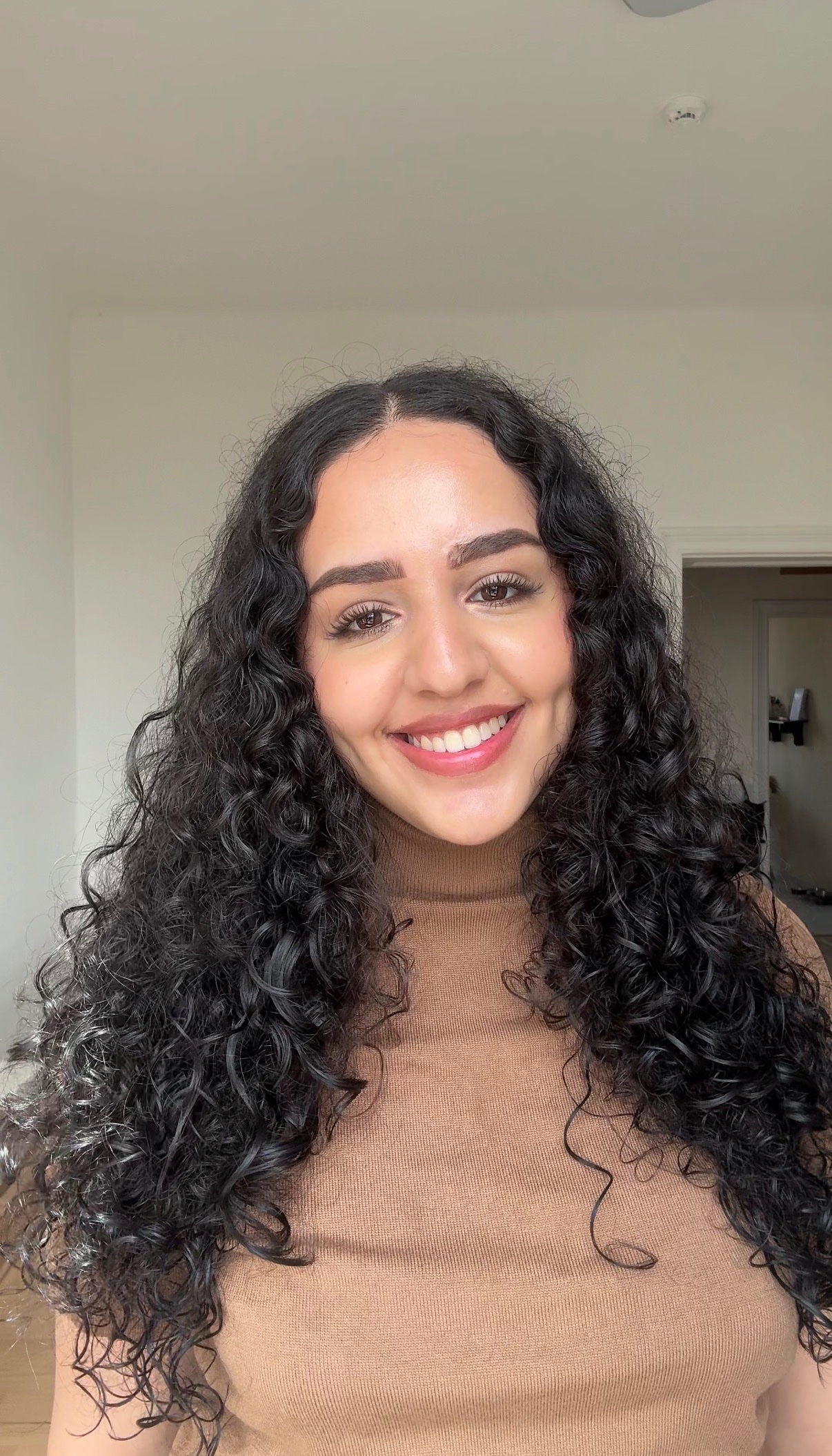 A young woman with curly hair smiles warmly at the camera, wearing a brown turtleneck sweater. The background is softly blurred, suggesting a cozy indoor setting.