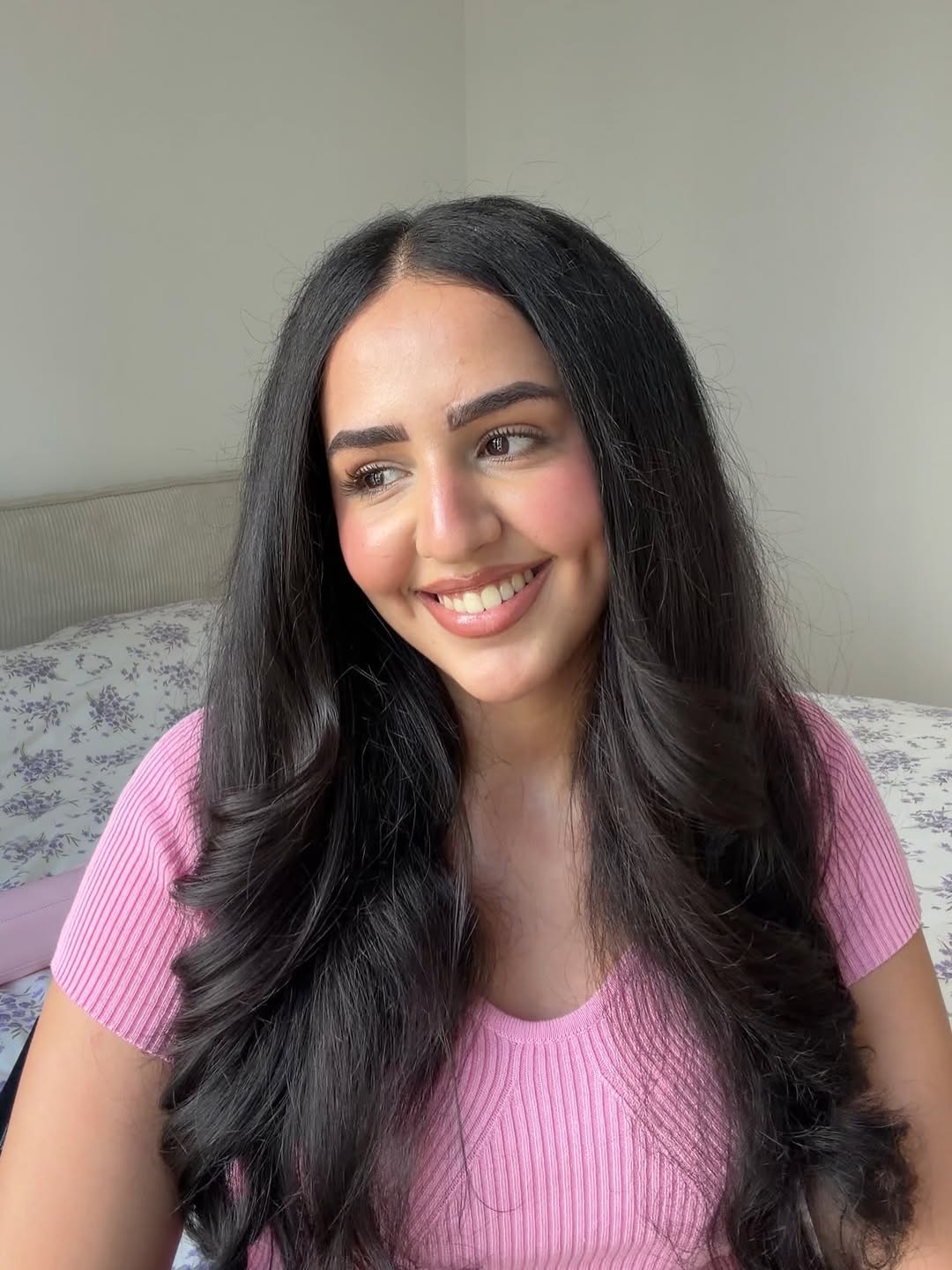 A young woman with long, wavy black hair smiles warmly while sitting on a bed with floral bedding.