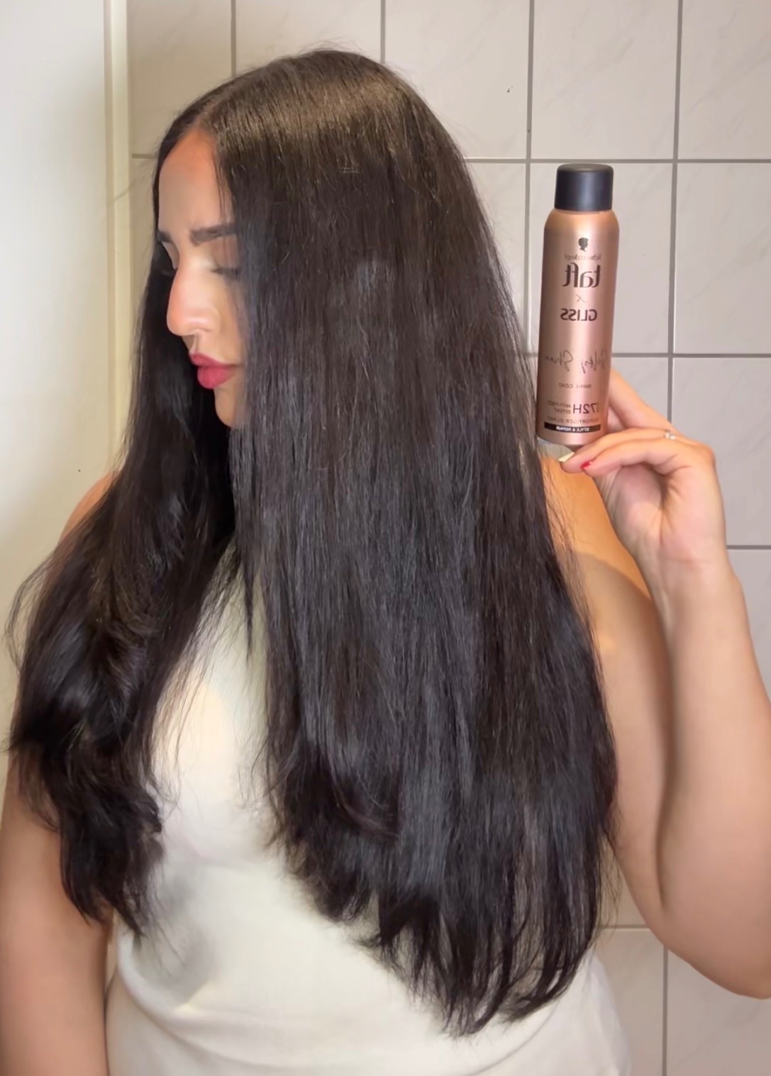 A woman with long, shiny hair holds a hair product bottle while looking to the side.
