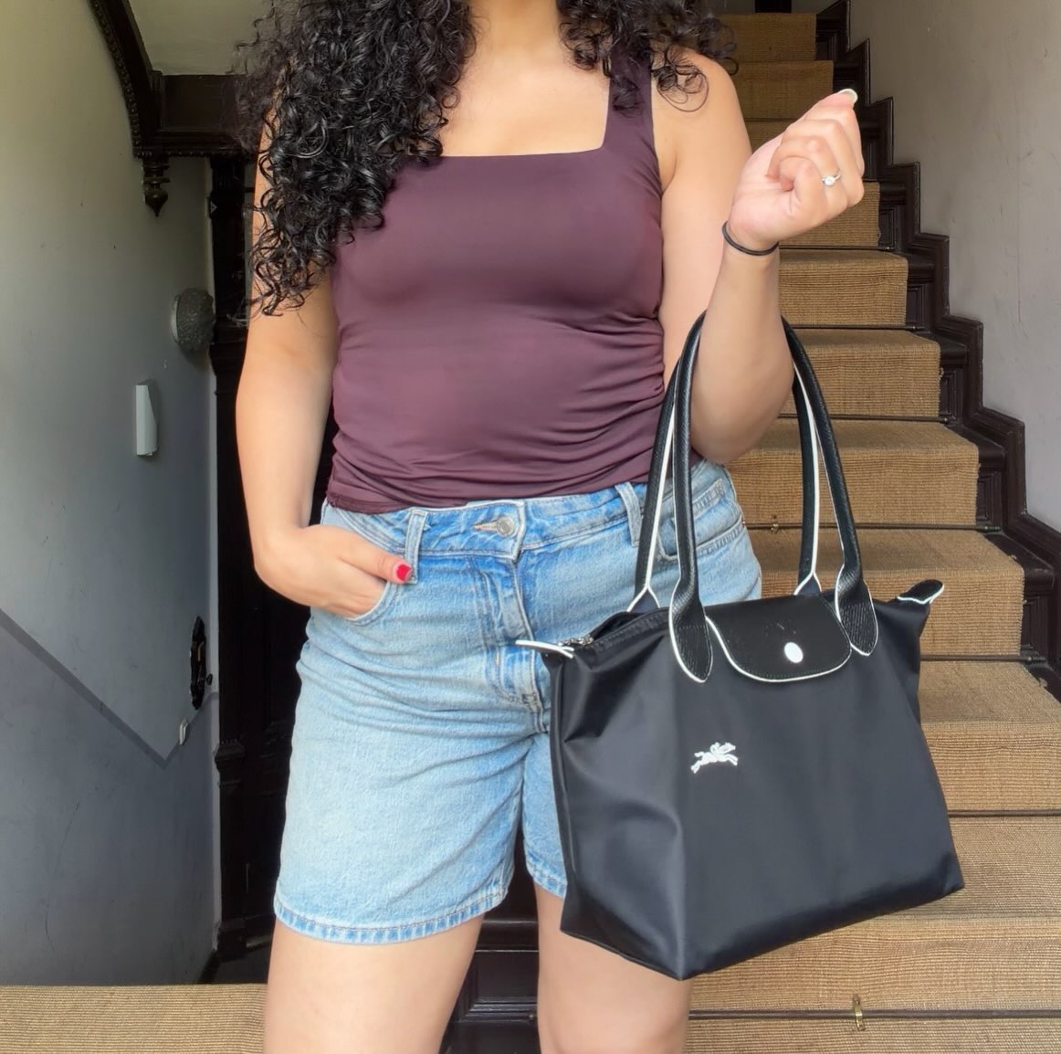 A person stands on a staircase, wearing a brown tank top and denim shorts, holding a black handbag.