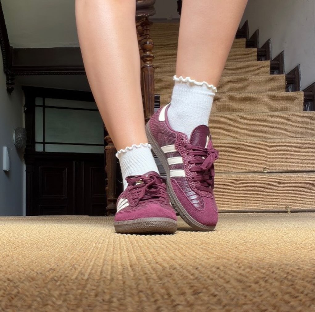 A close-up of a person's feet wearing burgundy sneakers and white socks, standing on a textured rug with wooden stairs in the background.