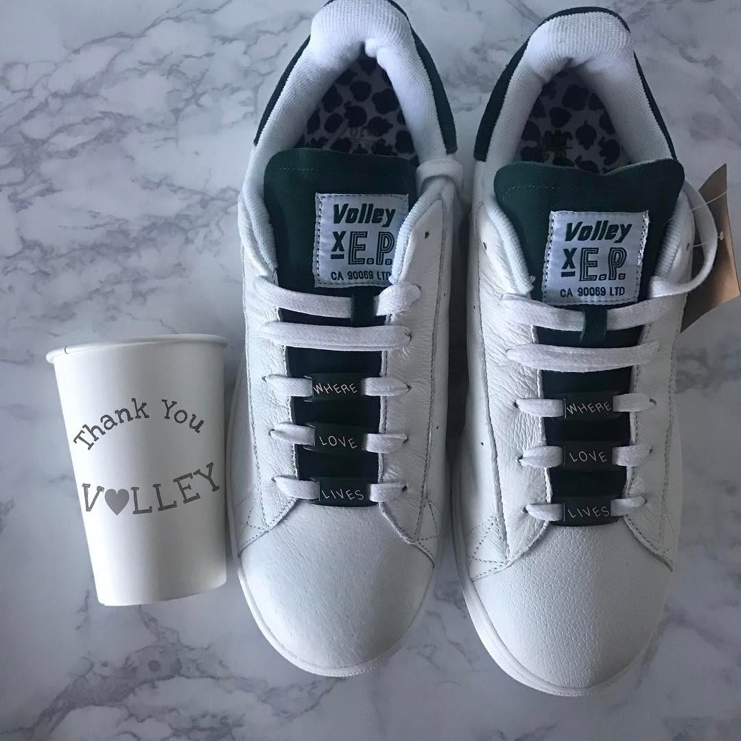 A pair of white sneakers with green accents placed next to a coffee cup that has the text 'Thank You VOLLEY' printed on it.