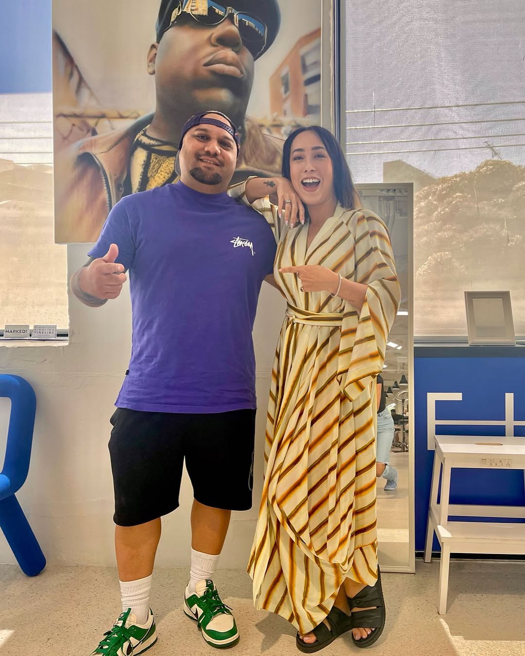 Two individuals pose together, smiling and giving thumbs up, in front of a large portrait of a rapper. One person wears a purple shirt and shorts, while the other is dressed in a striped, flowing dress.