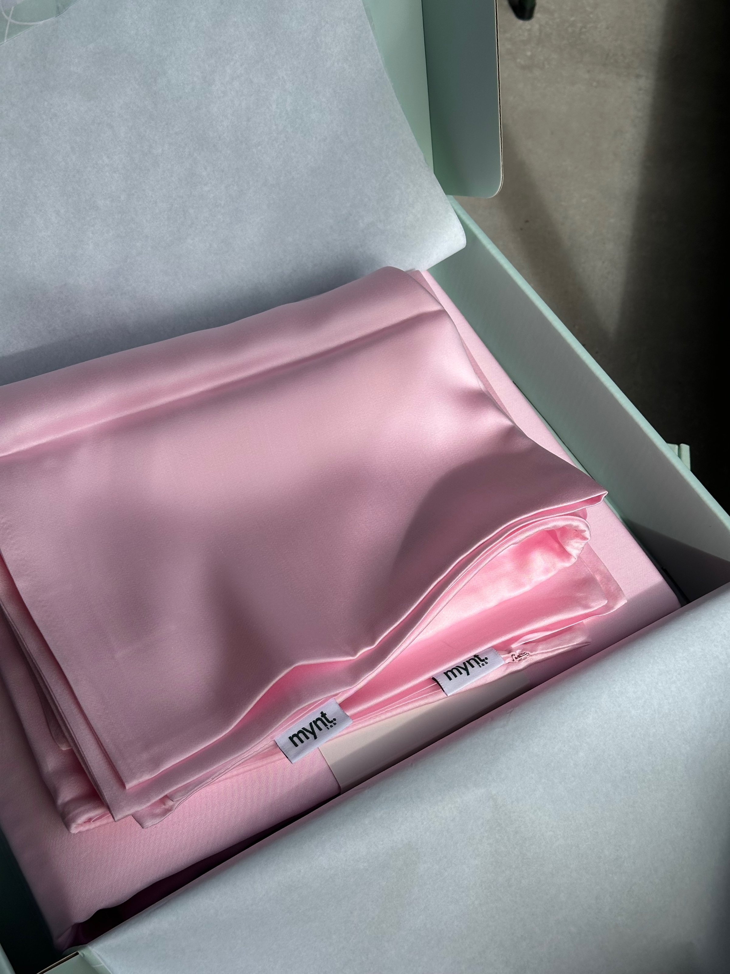 A neatly folded pink satin fabric placed inside a light green box, partially covered with tissue paper.