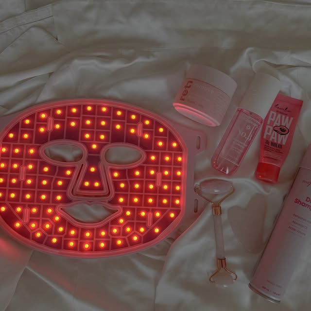 A skincare setup featuring a red light therapy mask, various skincare products, and a jade roller arranged on a white fabric background.
