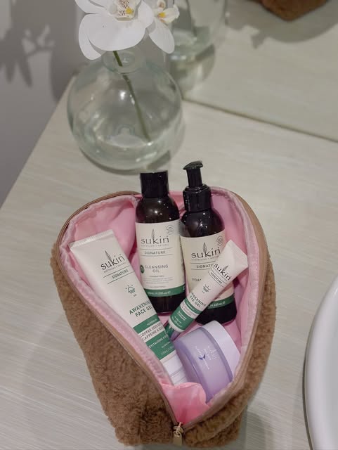 A heart-shaped cosmetic bag filled with various skincare products from Sukin, placed on a bathroom counter next to a glass vase with a flower.
