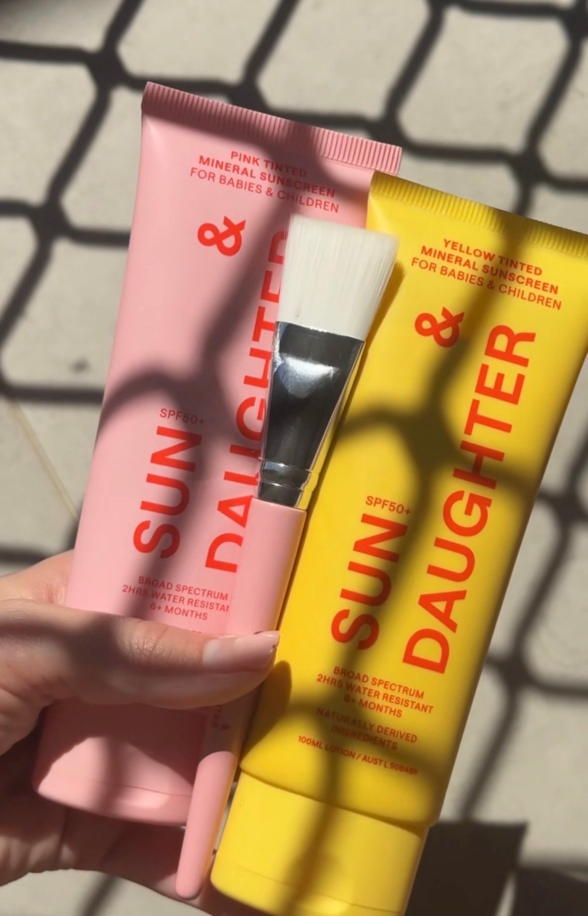 Two tubes of sunscreen for babies and children, one pink and one yellow, held in a hand with a brush beside them.