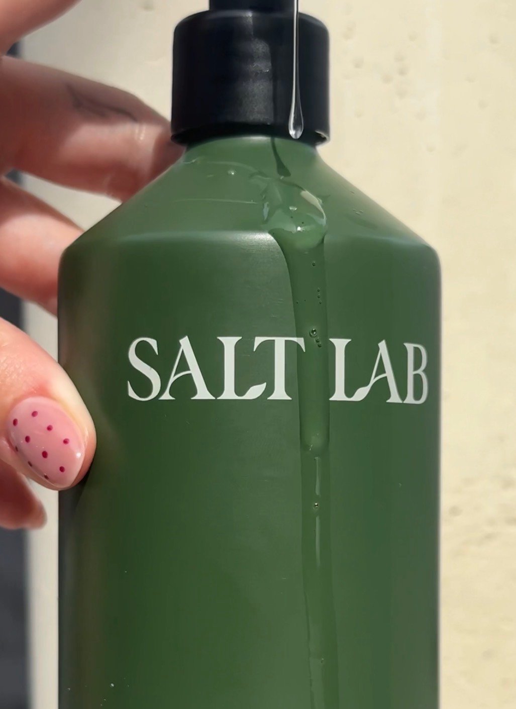 A close-up of a green bottle labeled 'SALT LAB' with liquid dripping down the side, held by a hand with pink nail polish featuring polka dots.