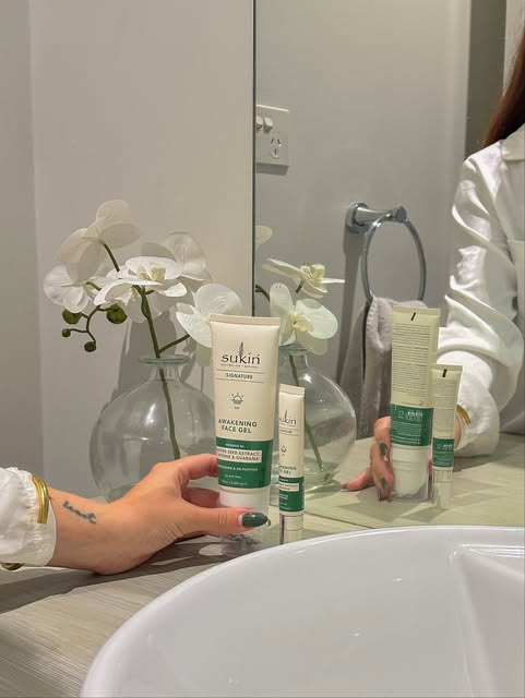 A hand holding Sukin skincare products in front of a mirror, with a vase of white orchids in the background.