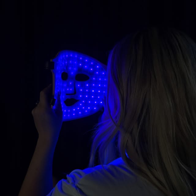 A person with long hair holds a blue LED mask against their face, set against a dark background.