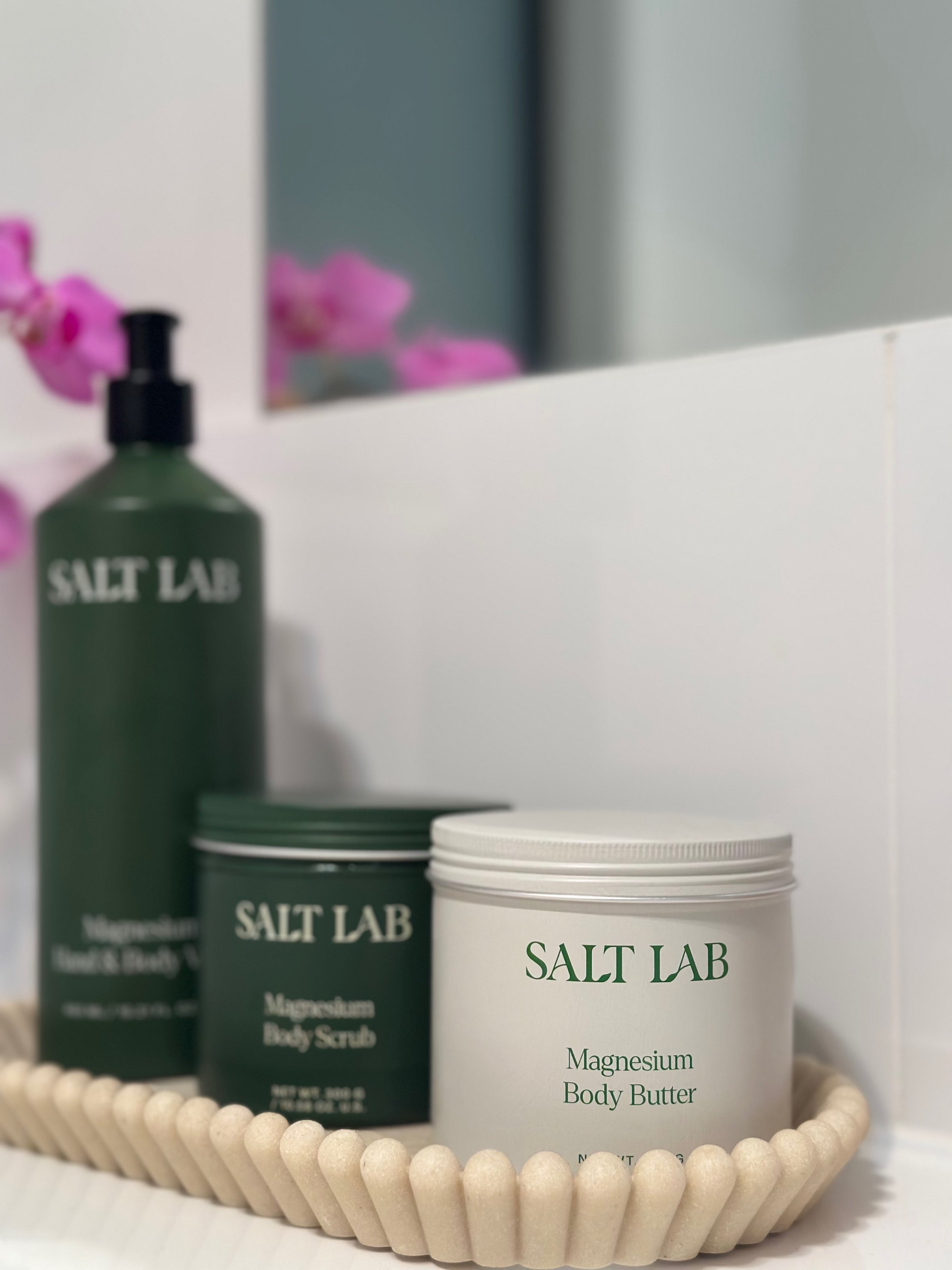 A collection of SALT LAB skincare products displayed on a textured tray, featuring a body scrub, body butter, and body wash, with pink orchids in the background.