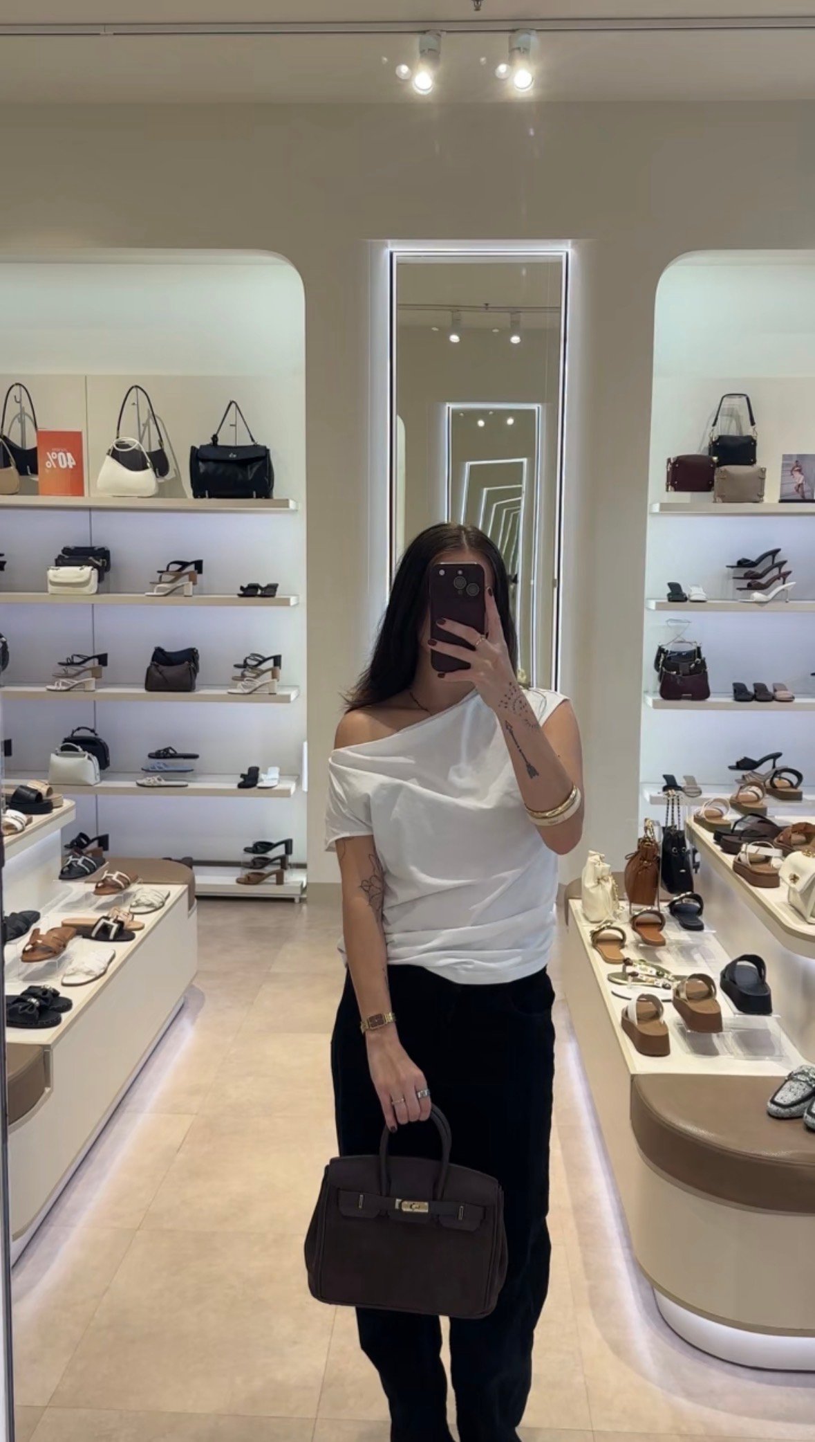 A person stands in a stylish store holding a brown handbag, surrounded by shelves displaying various shoes and bags.