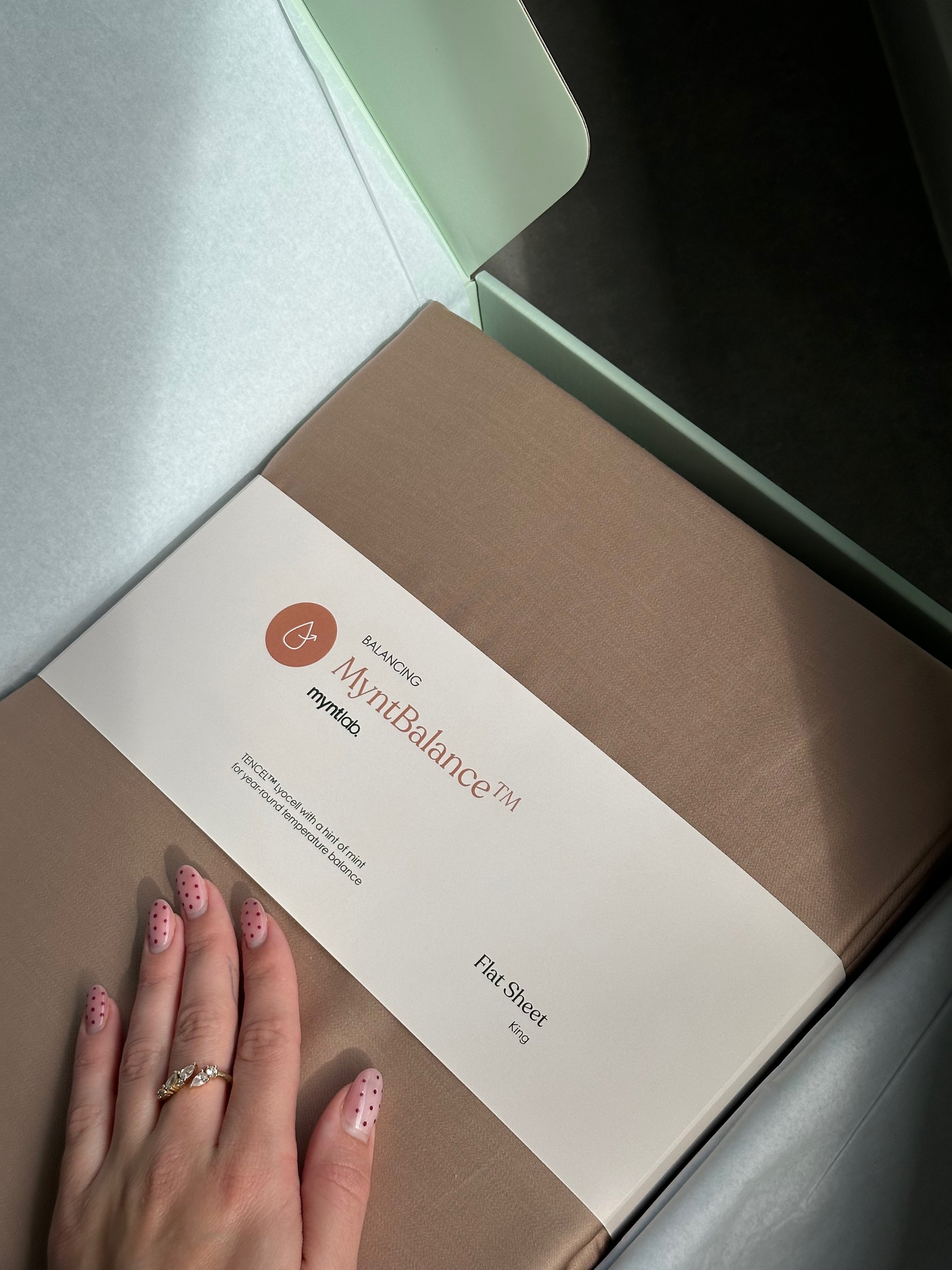 A hand with pink polka-dotted nails rests on a brown package labeled 'MyNutraBalance TAI' inside a light green box.