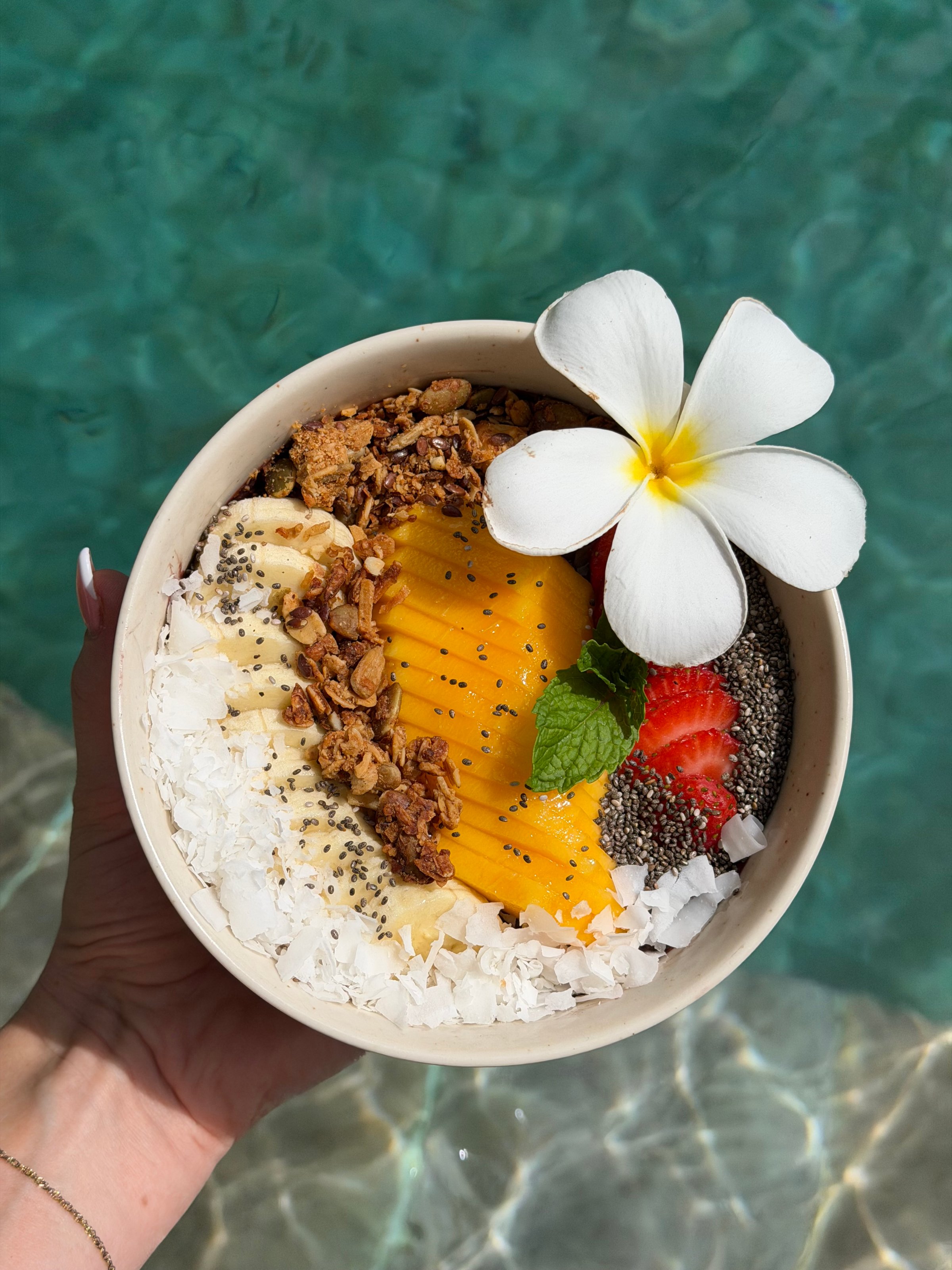 A colorful smoothie bowl topped with mango, banana, strawberries, granola, chia seeds, coconut flakes, and a flower, held above clear blue water.