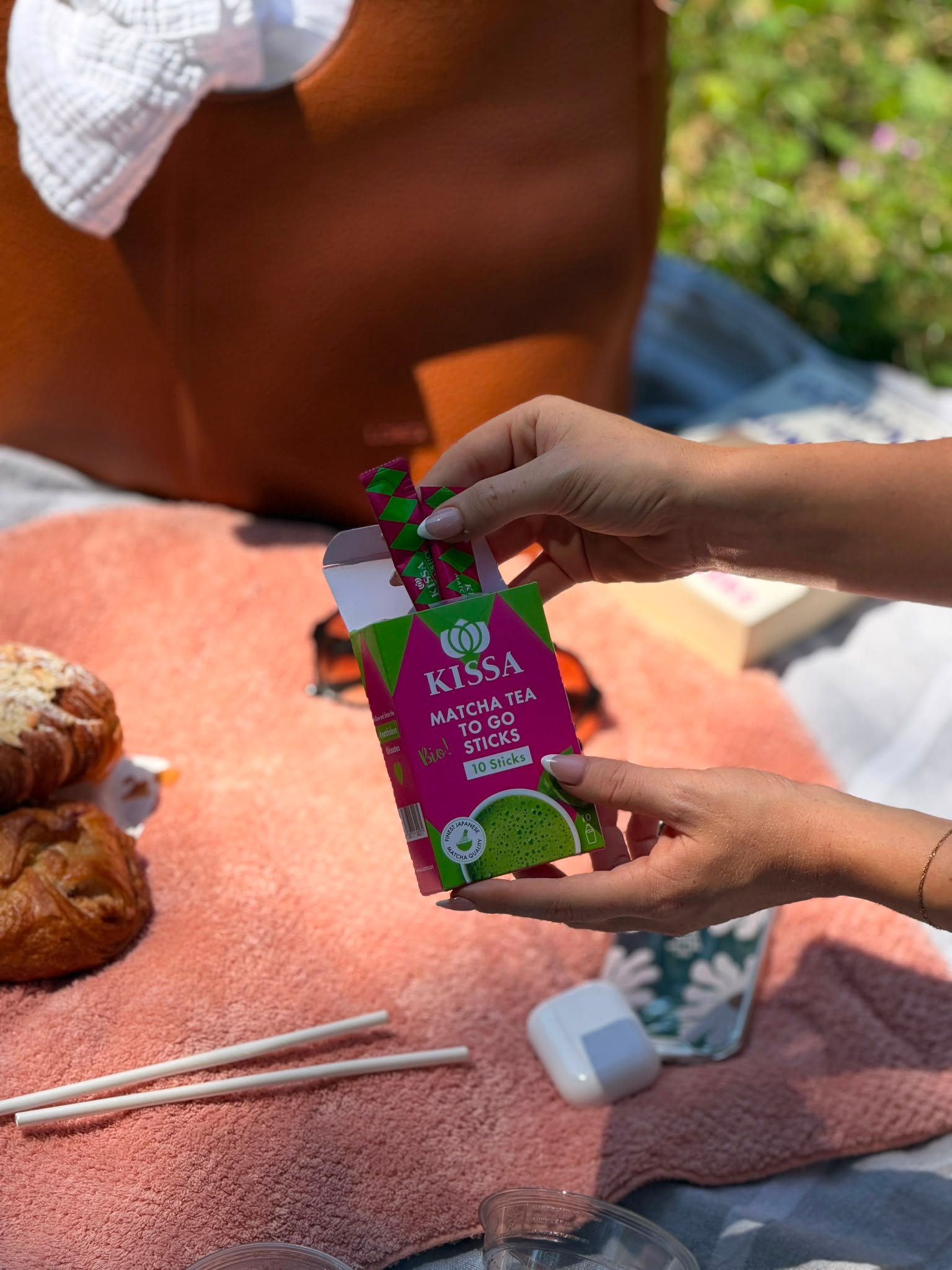 A hand holding a box of Kissa matcha tea sticks over a picnic setup with pastries and chopsticks on a pink cloth.