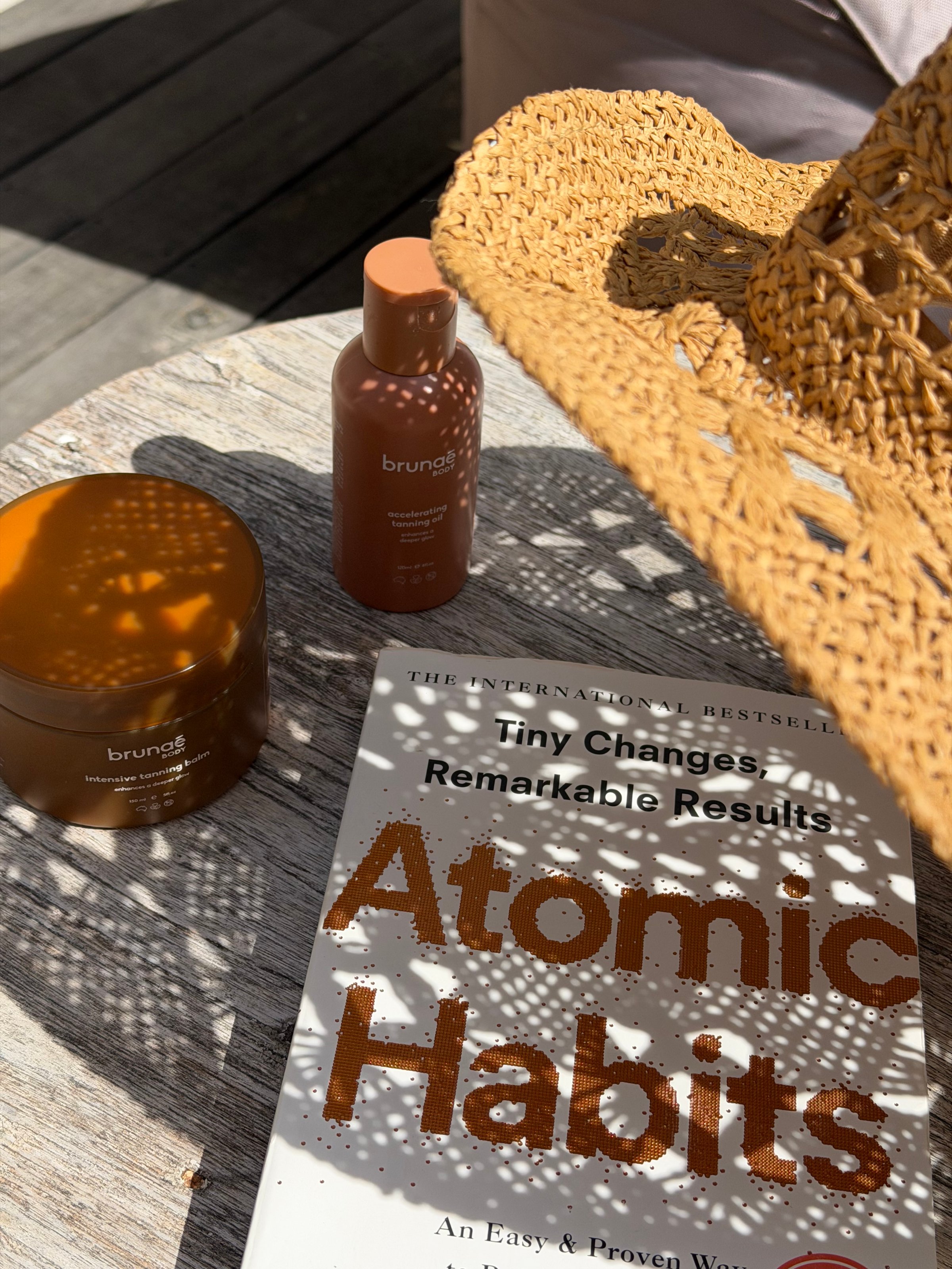 A sunlit outdoor scene featuring a straw hat, skincare products, and the book 'Atomic Habits' on a wooden table.