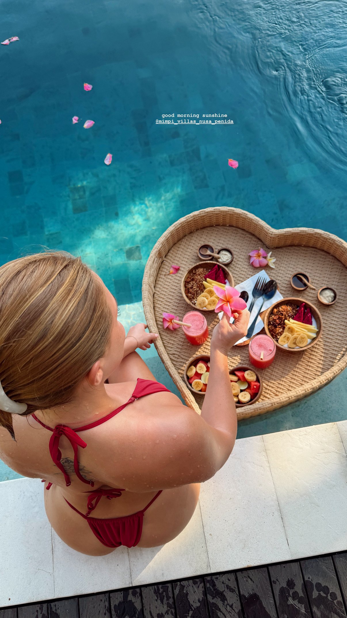 A woman in a red swimsuit enjoys a heart-shaped breakfast tray by a pool, surrounded by flower petals.