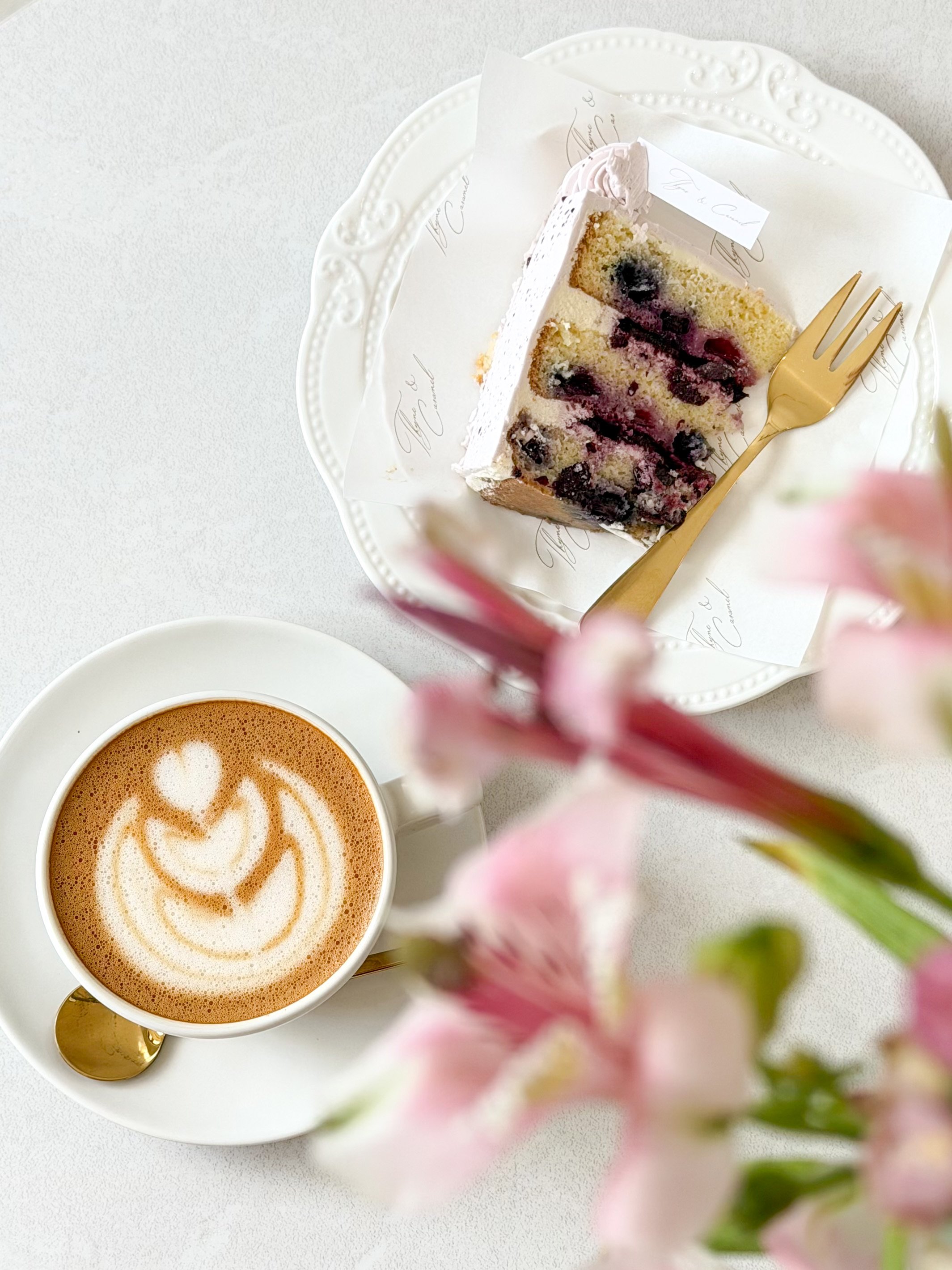 A slice of blueberry cake on a decorative plate next to a cup of latte with a heart design in the foam, surrounded by pink flowers.
