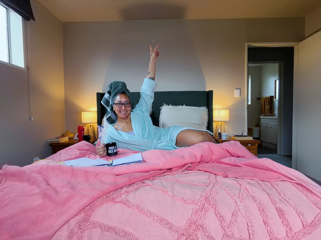 A person in a light blue robe and towel on their head is playfully posing on a pink bed, with a coffee mug in hand and a cheerful expression.