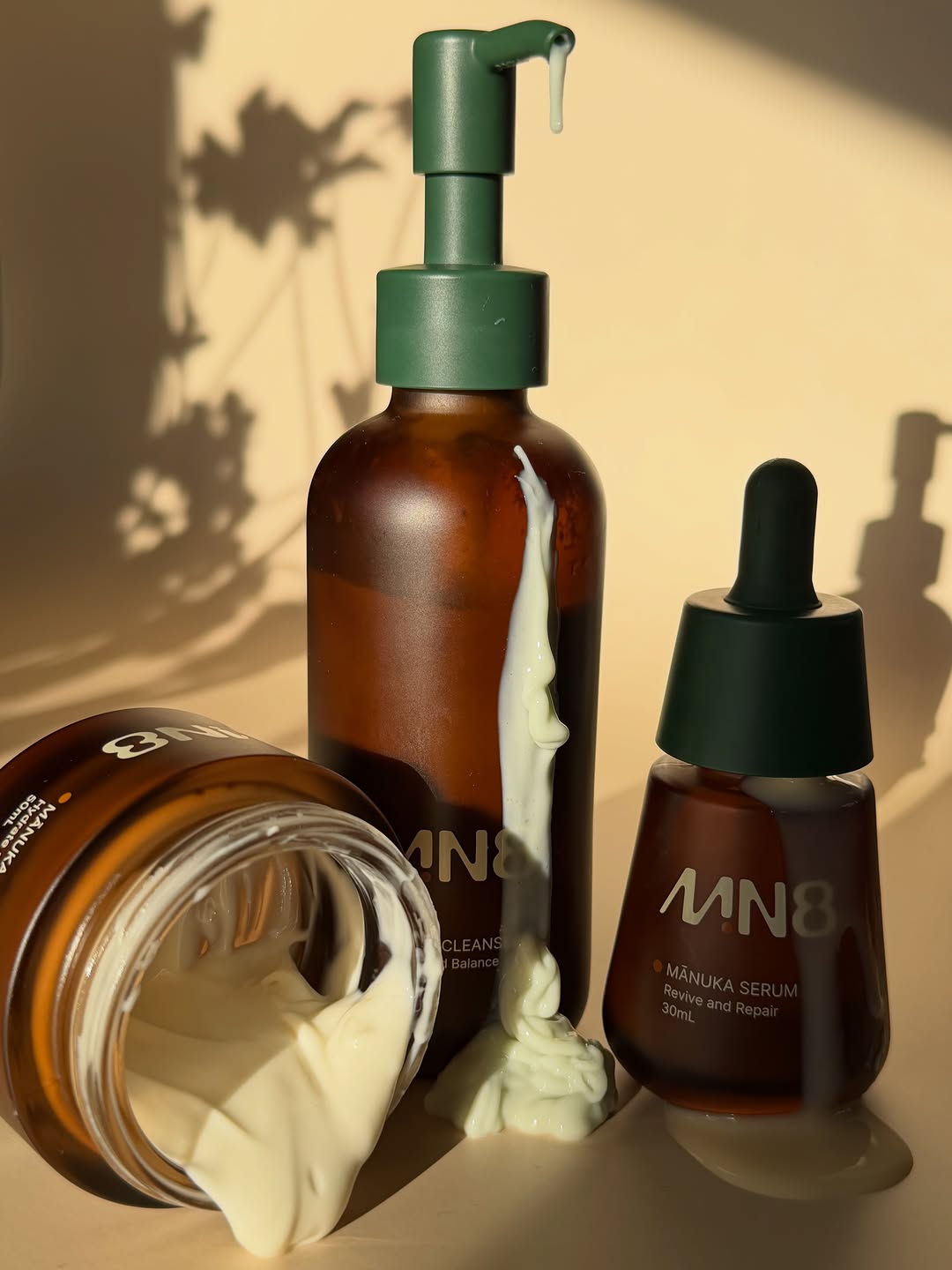 Three skincare products with a creamy texture displayed against a warm, shadowy background. The products include a cleanser and a serum, showcasing their sleek packaging.