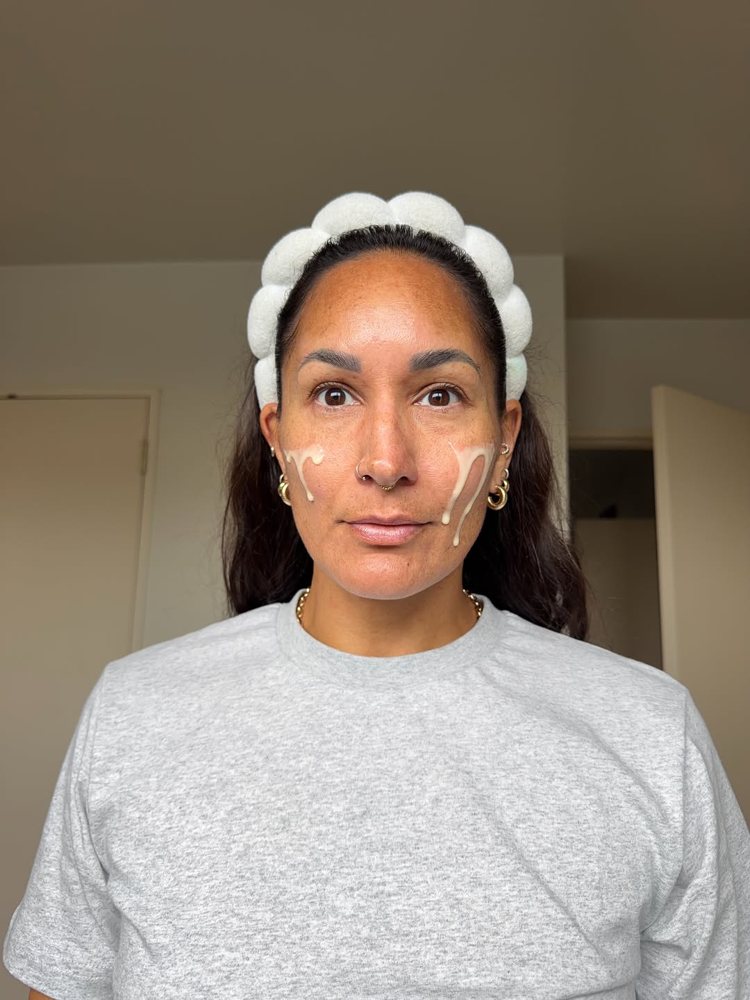 A person with a headband and skincare product applied to their face, standing in a well-lit indoor space.