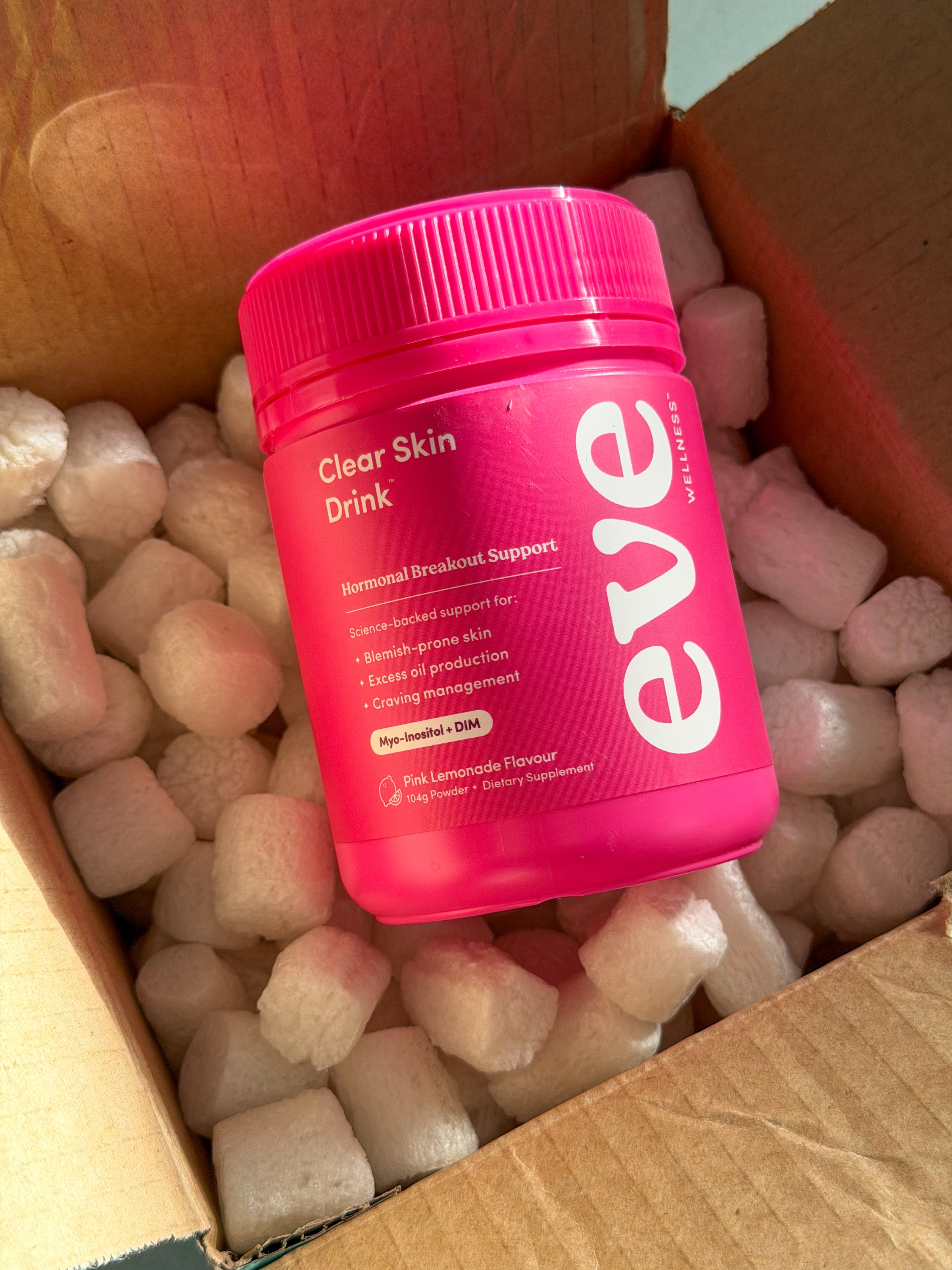 A bright pink container labeled 'Clear Skin Drink' sits atop packing peanuts inside a cardboard box, suggesting a product delivery.