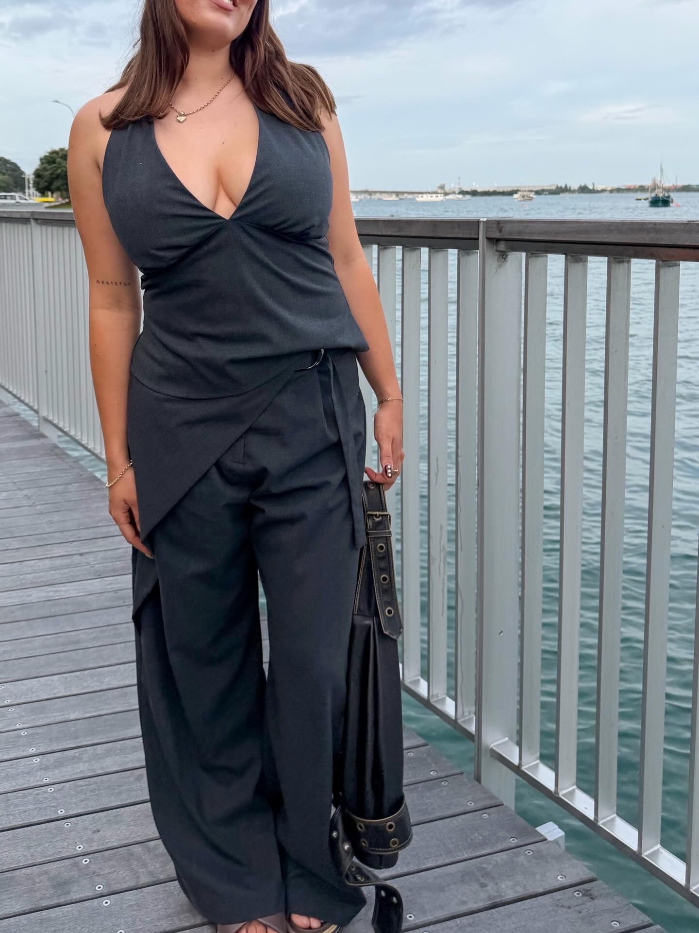 A woman stands on a wooden walkway by the water, wearing a stylish black outfit with a halter top and wide-leg pants.