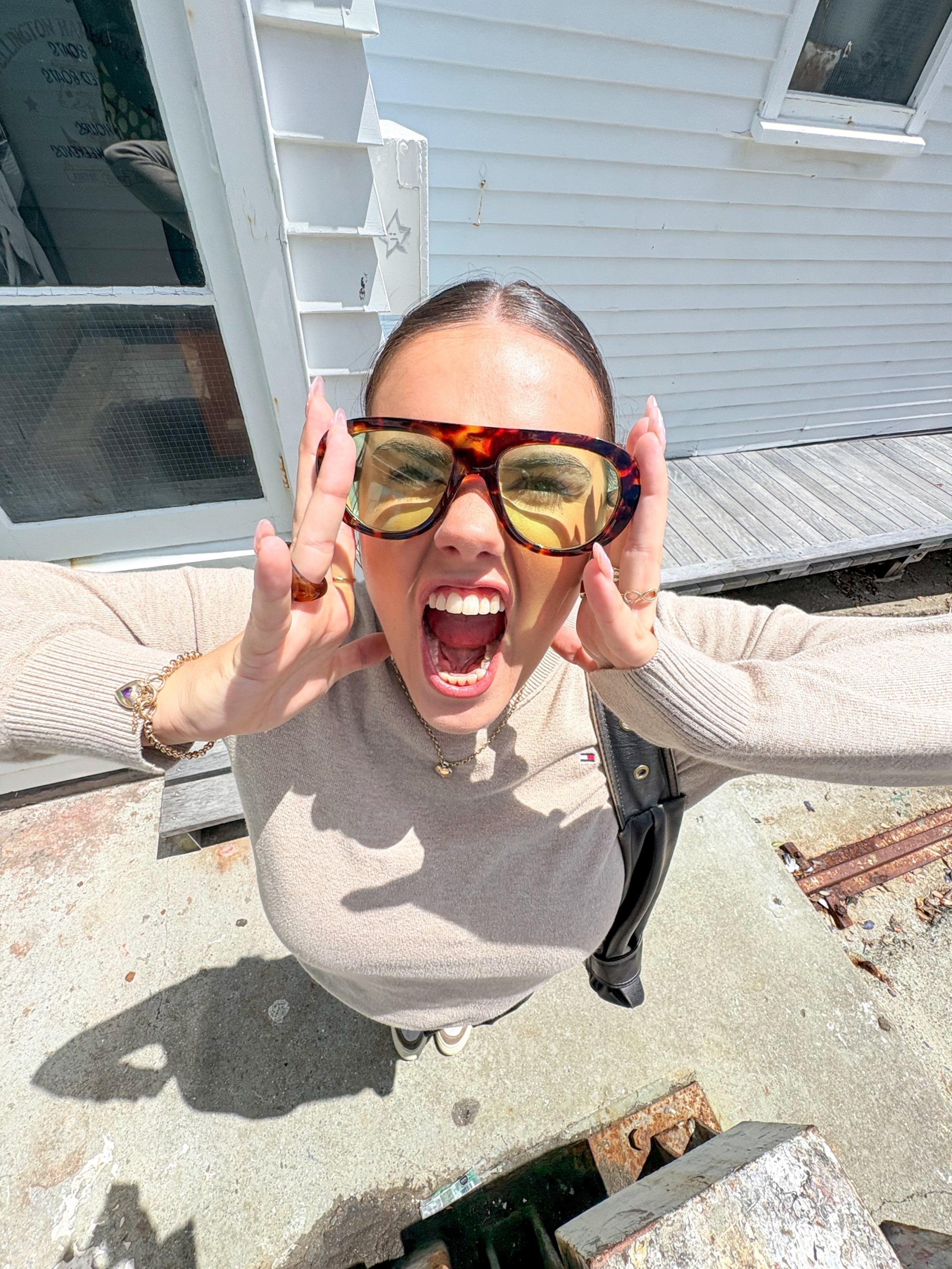A woman with long hair wearing oversized sunglasses and a beige sweater, expressing excitement while posing outdoors.