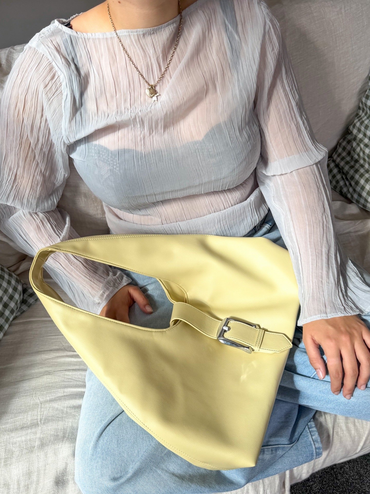 A person wearing a light blue textured top and denim jeans holds a yellow handbag, sitting on a couch with a checkered background.