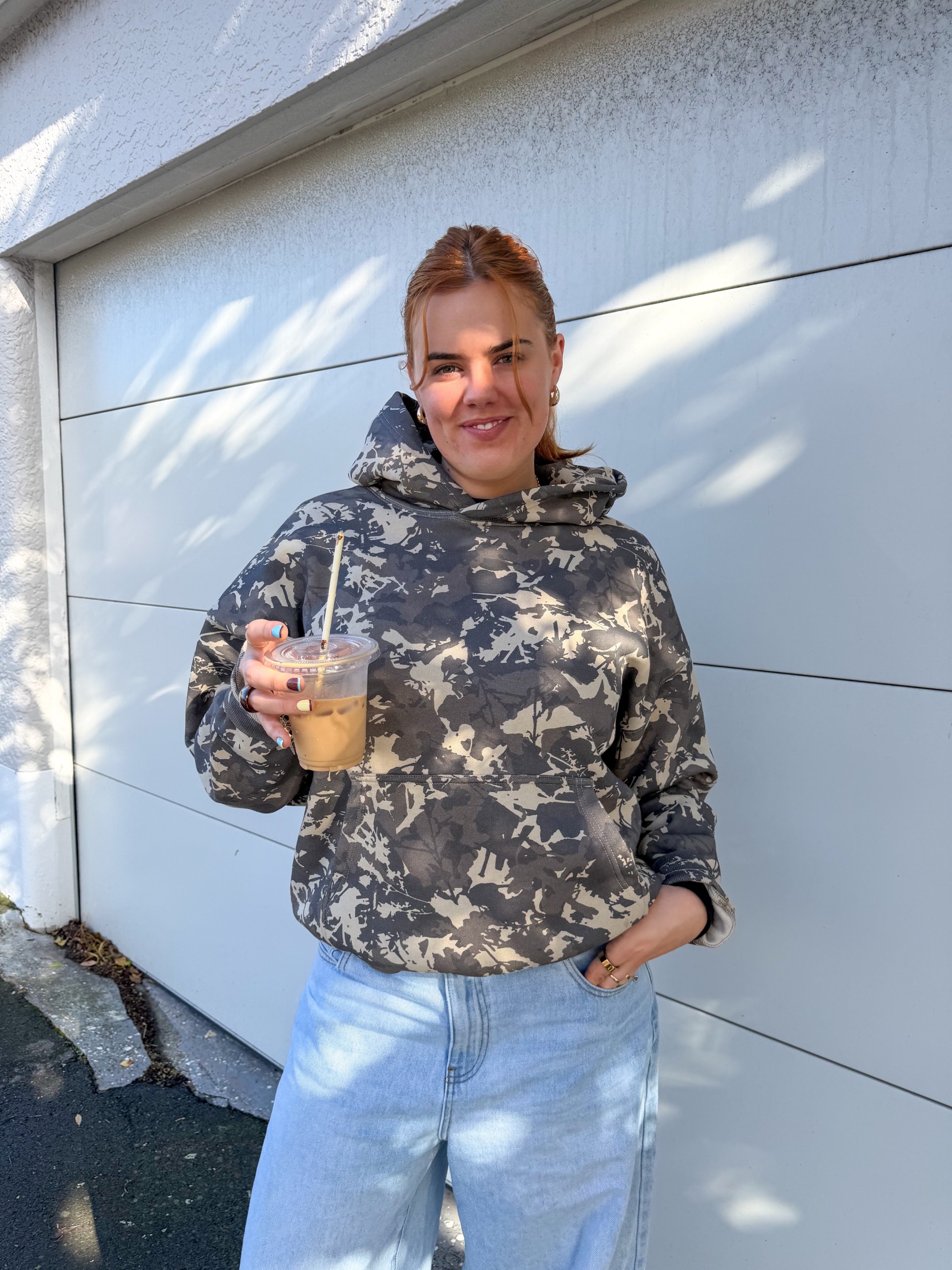 A person wearing a camouflage hoodie and light blue jeans stands outside, holding a cup of iced coffee with a smile.