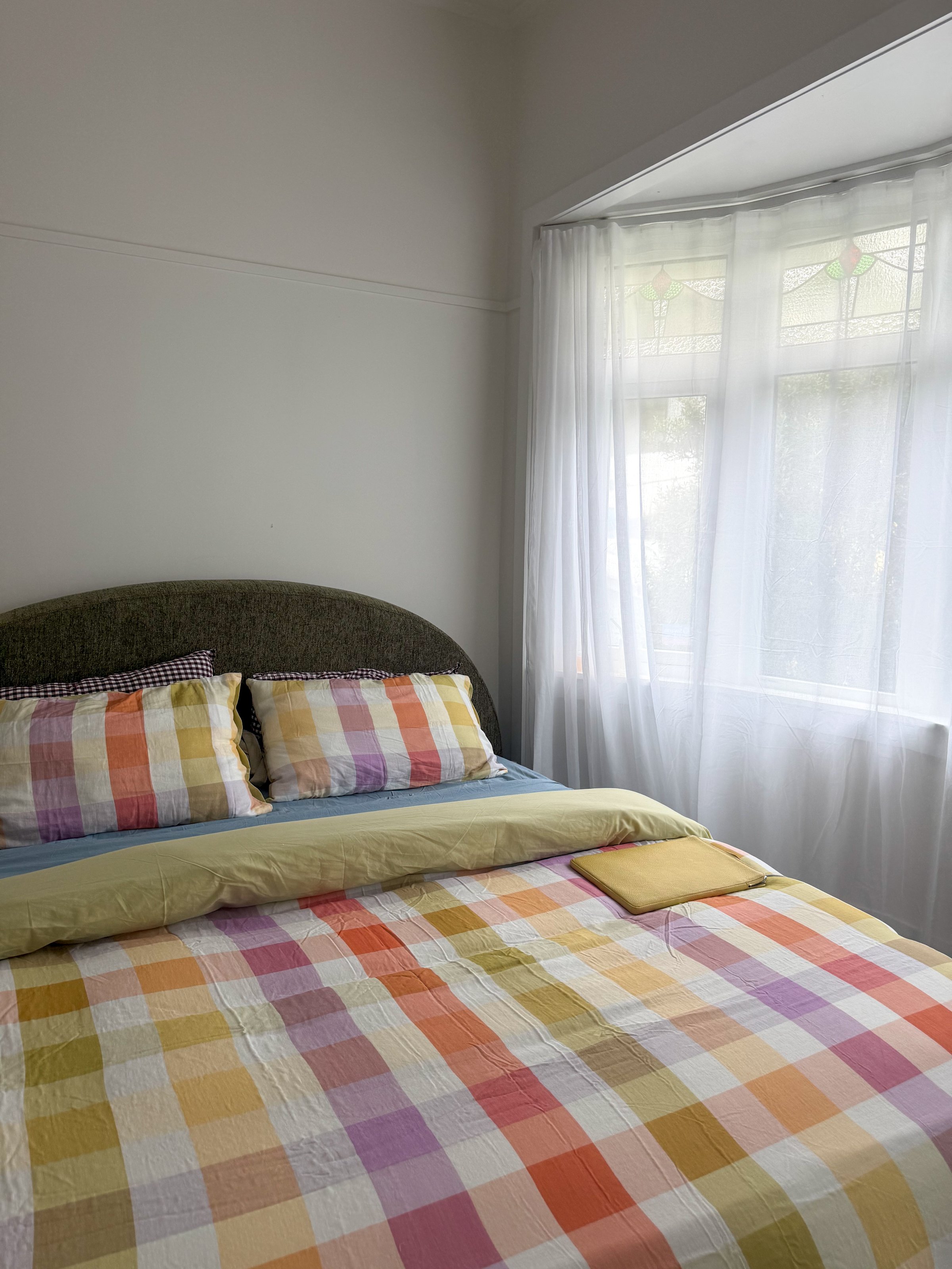 A cozy bedroom featuring a bed with a colorful plaid quilt and pillows, illuminated by natural light from a window with sheer curtains.