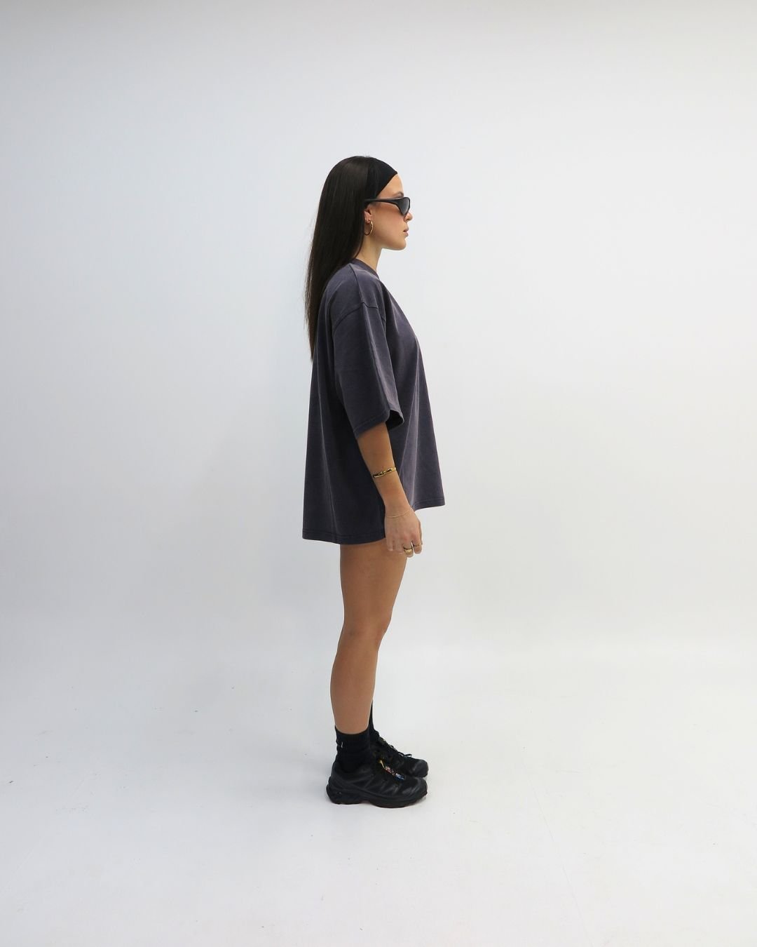 A person stands in profile wearing a loose-fitting gray t-shirt and sunglasses, showcasing a casual streetwear style.