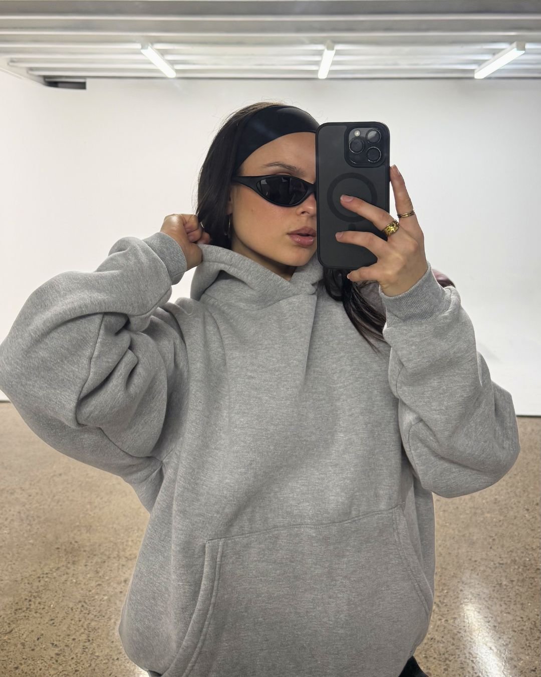 A person wearing a gray hoodie and sunglasses poses for a selfie in a minimalist studio setting.