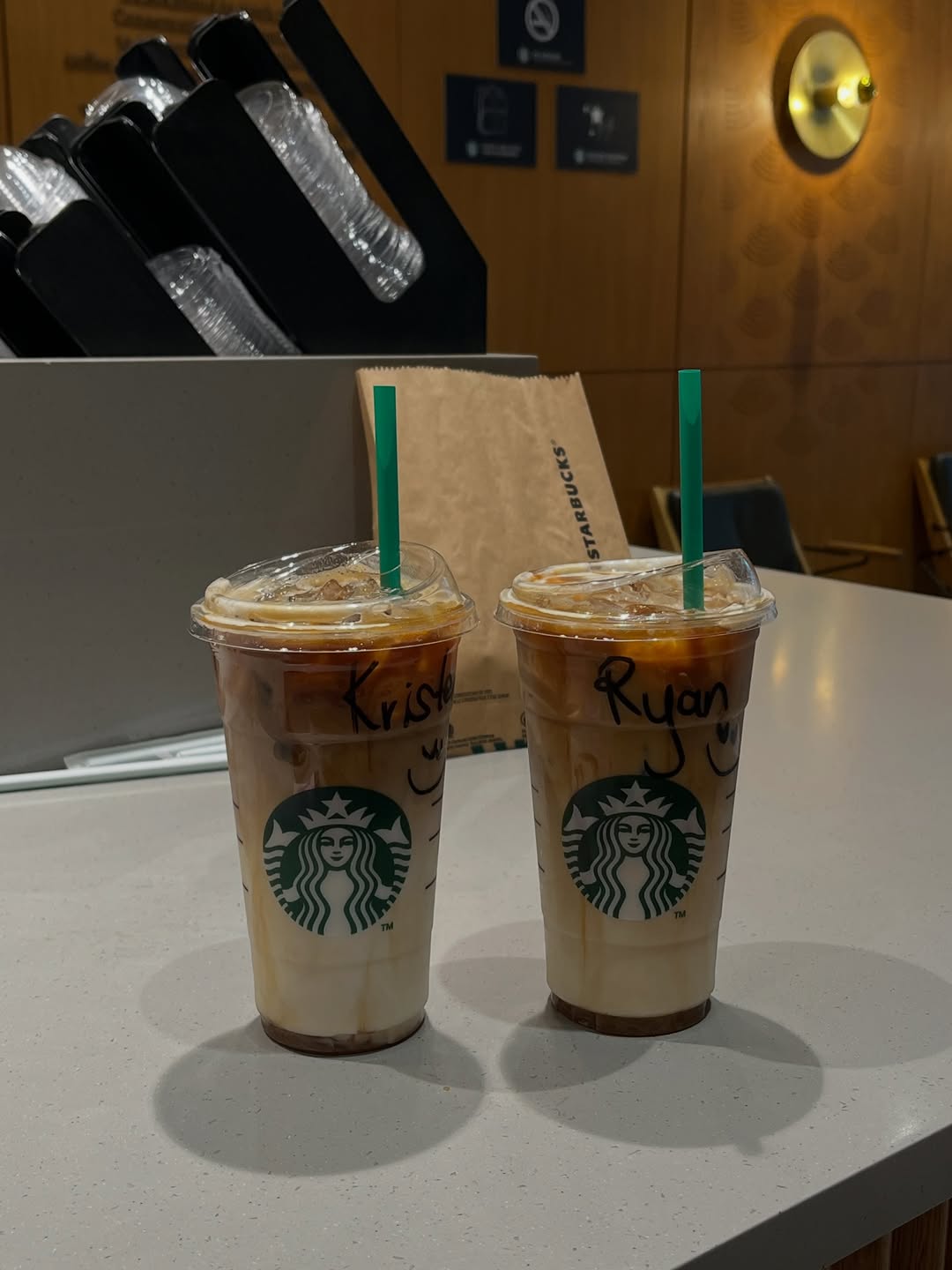Two iced coffee drinks with names written on them, placed on a table in a coffee shop.
