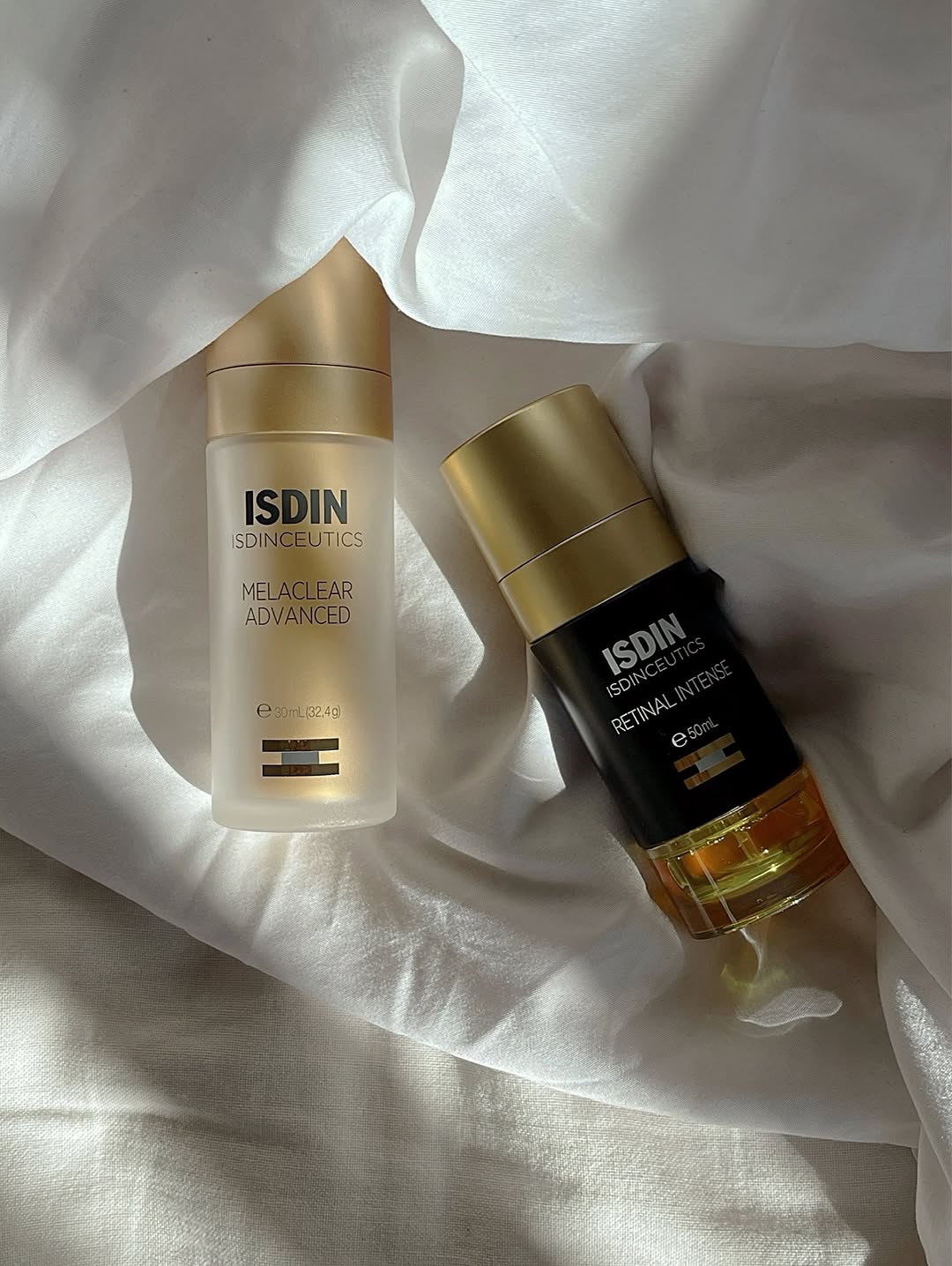 Two skincare products from ISDIN displayed on a soft, light-colored fabric. One is a serum and the other is an oil, both featuring elegant packaging.