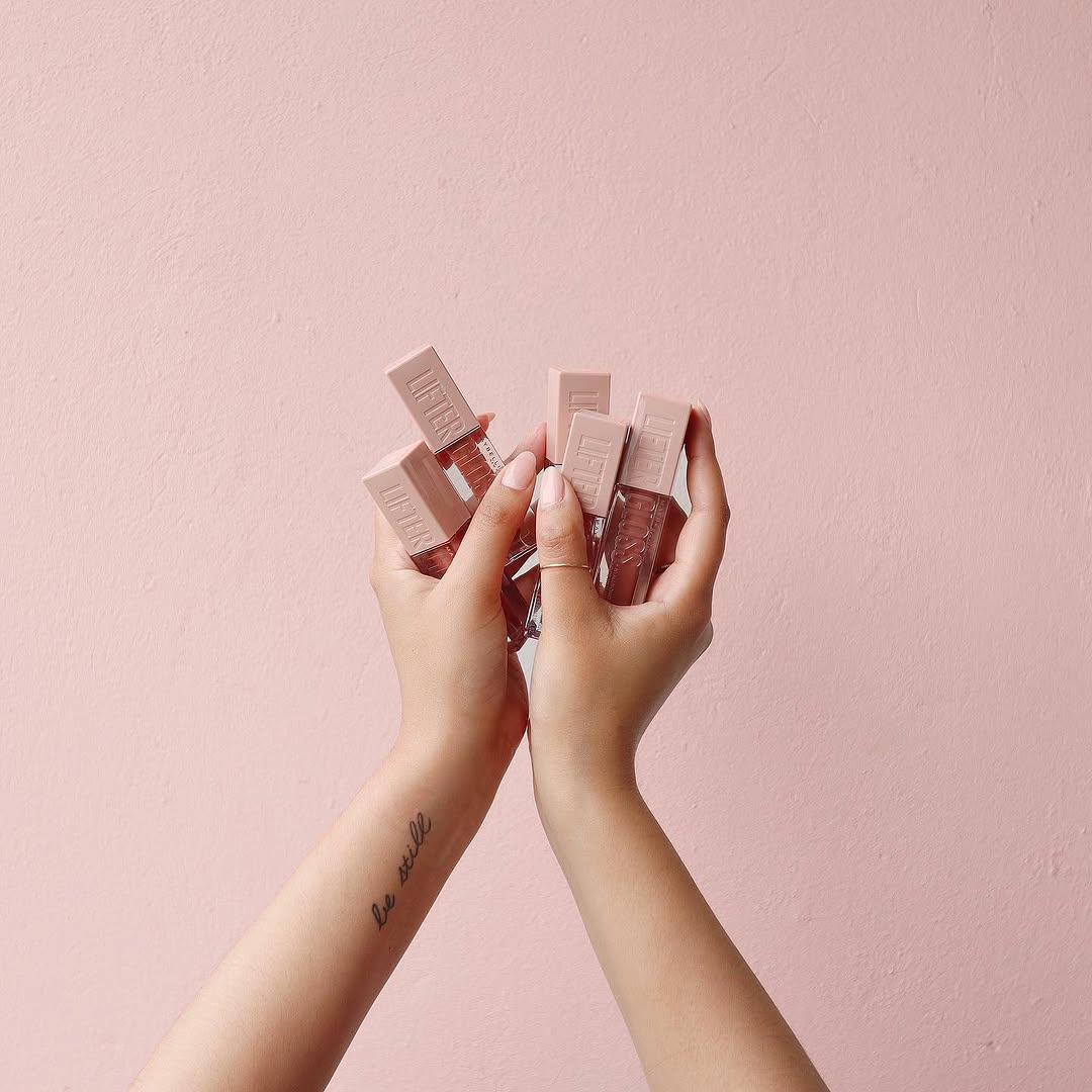 Two hands holding several bottles against a soft pink background, showcasing a beauty product collection.