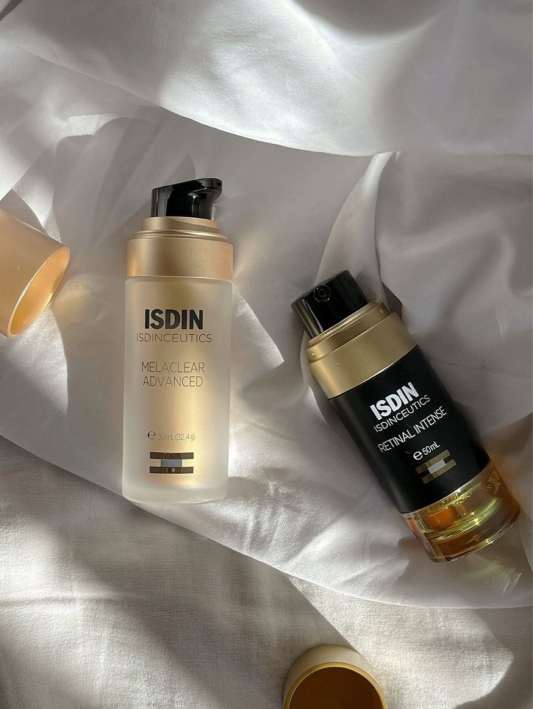 Two skincare products from ISDIN displayed on a soft, white fabric background. One is a clear bottle labeled 'Melaclear Advanced' and the other is a dark bottle labeled 'Retinal Intense'.
