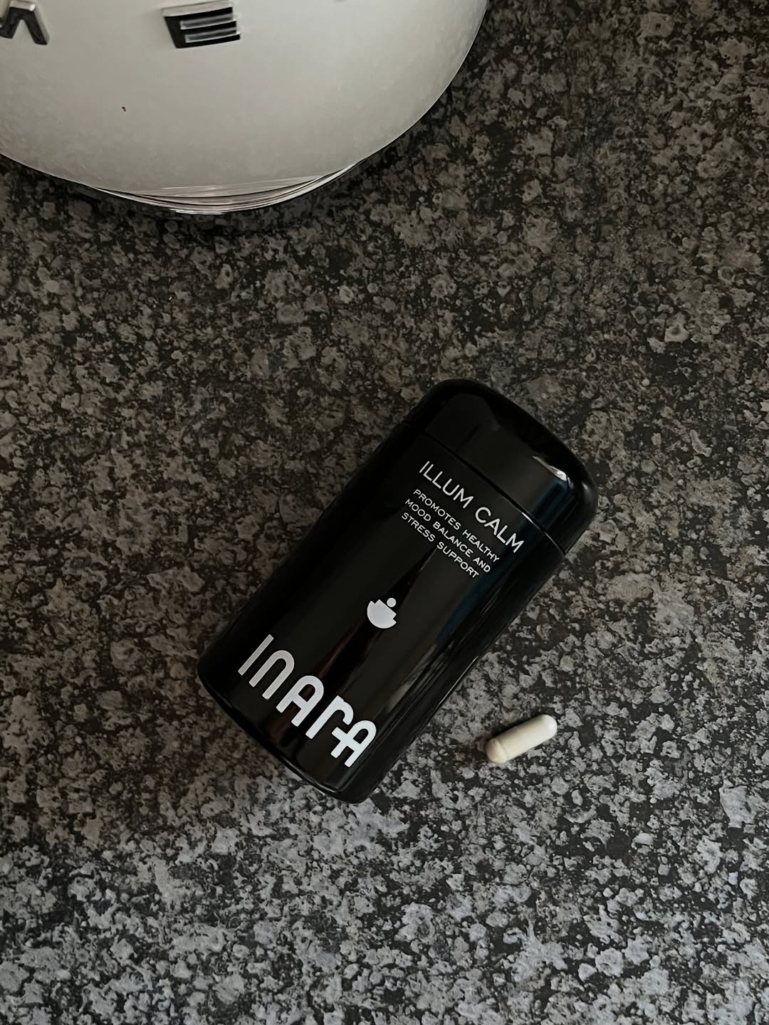A black cylindrical container labeled 'ILLUM CALM' sits on a textured gray surface, accompanied by a small white capsule.