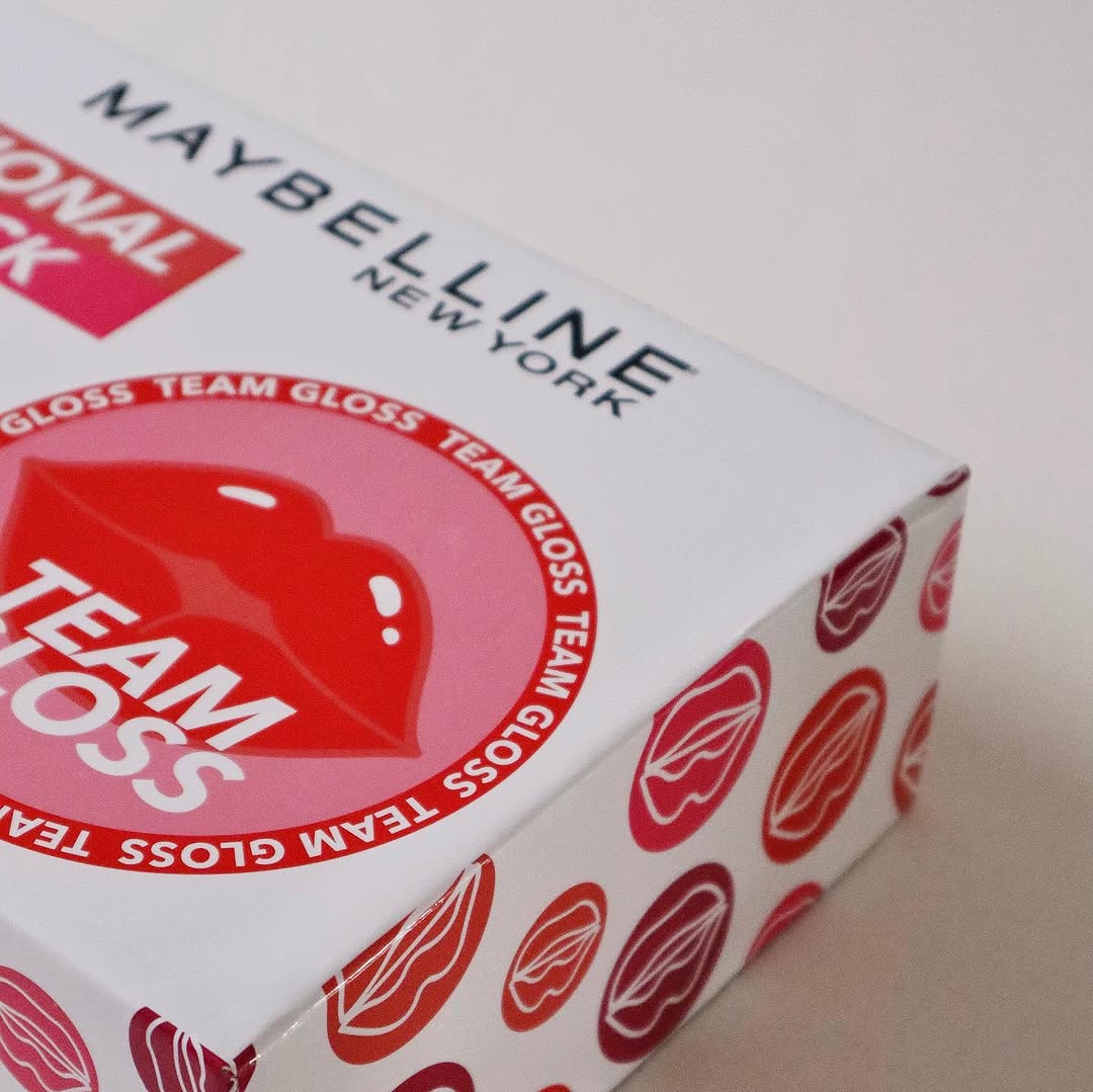 A close-up of a Maybelline product box featuring the Team Gloss branding and a lip print design.