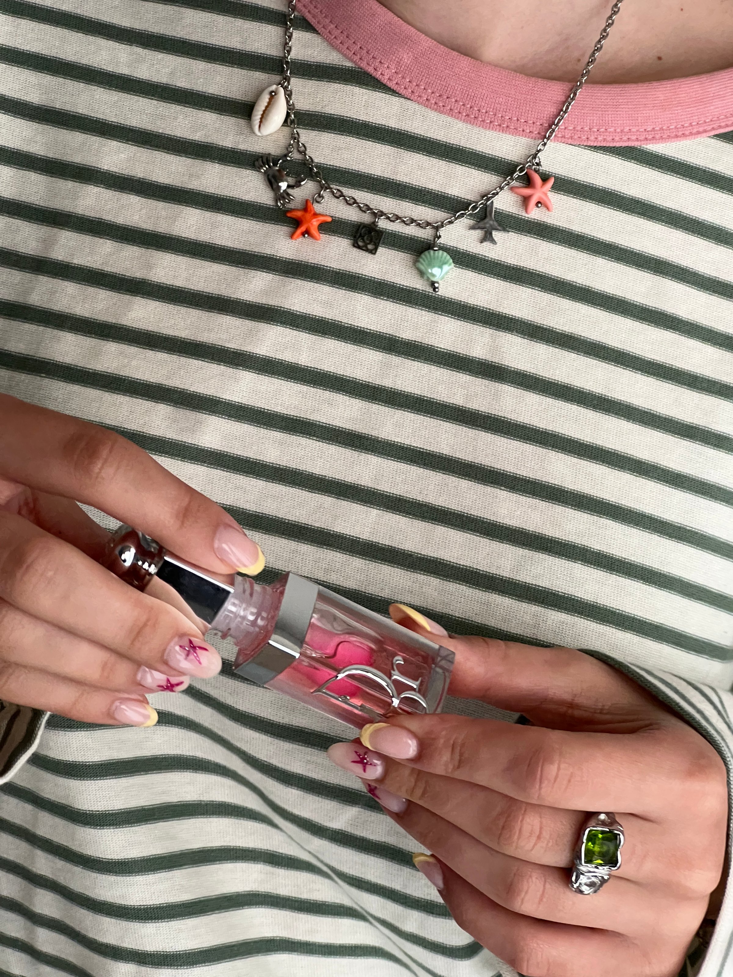 A person holding a bottle of nail polish with a pink hue, wearing a striped shirt and a charm necklace featuring starfish and seashells.