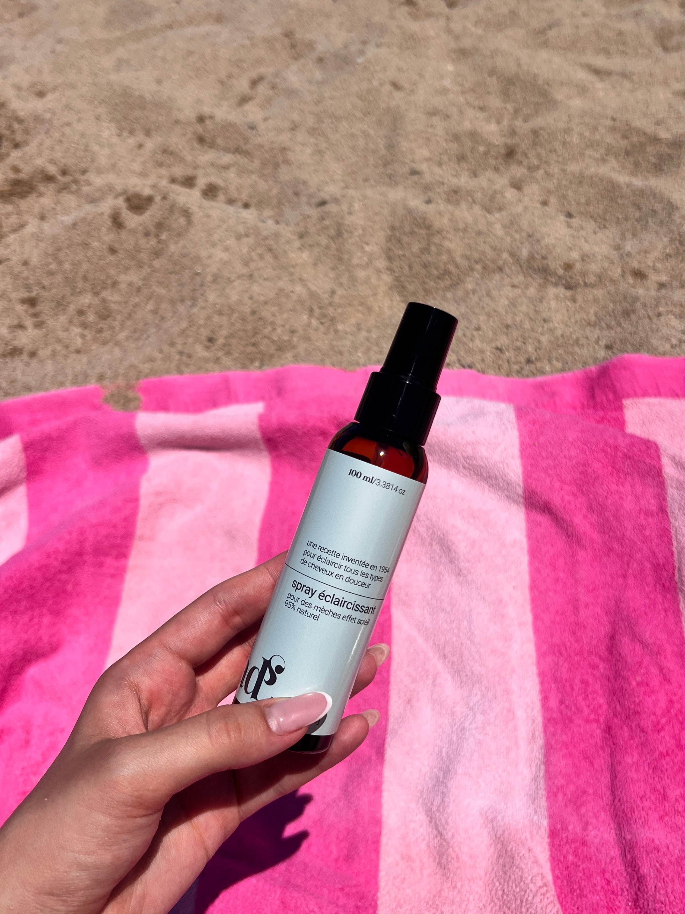 A hand holding a spray bottle of body moisturizer on a pink striped beach towel.