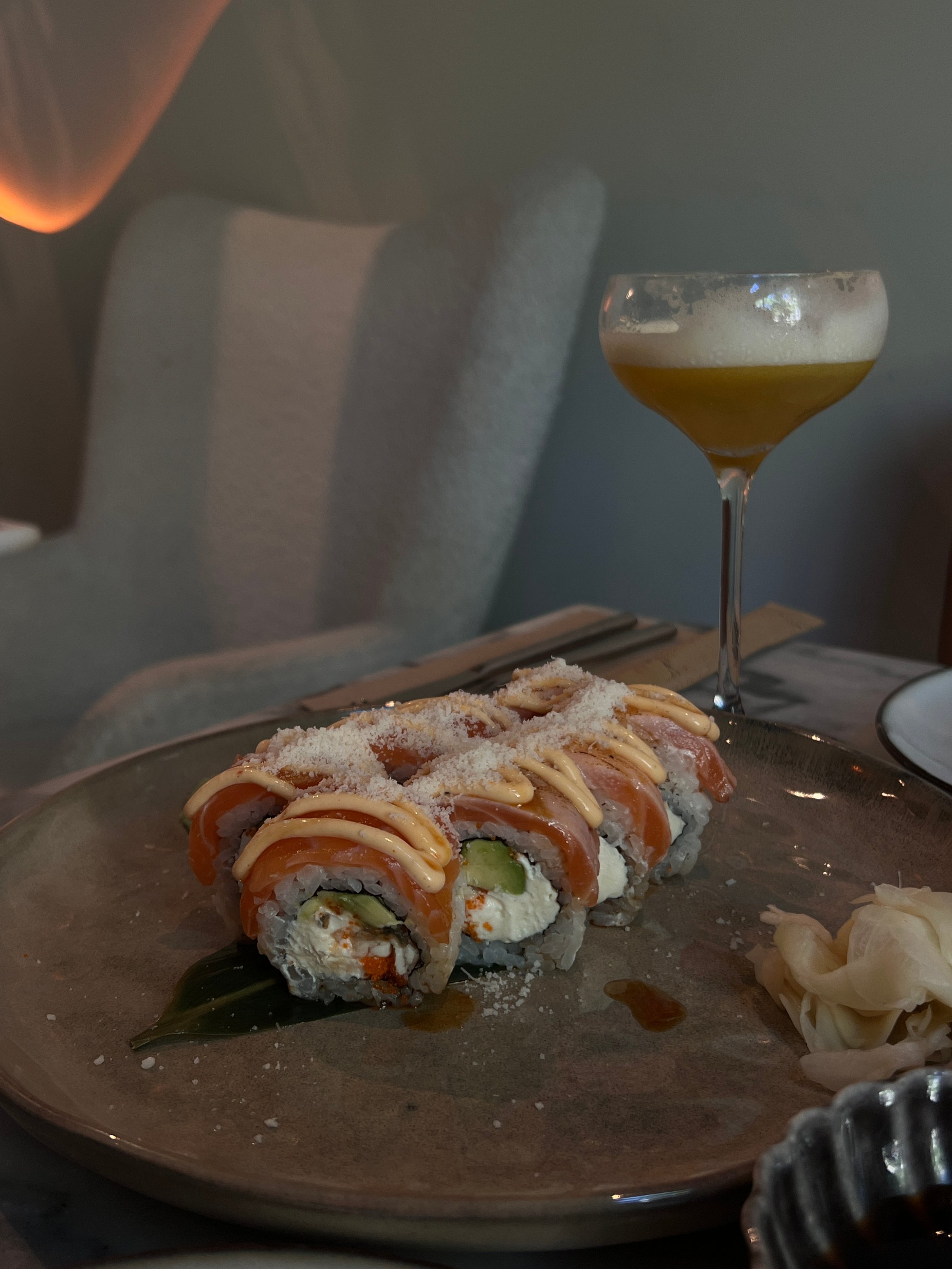 A beautifully arranged plate of sushi rolls topped with salmon and a drizzle of sauce, accompanied by pickled ginger and a cocktail in a glass.