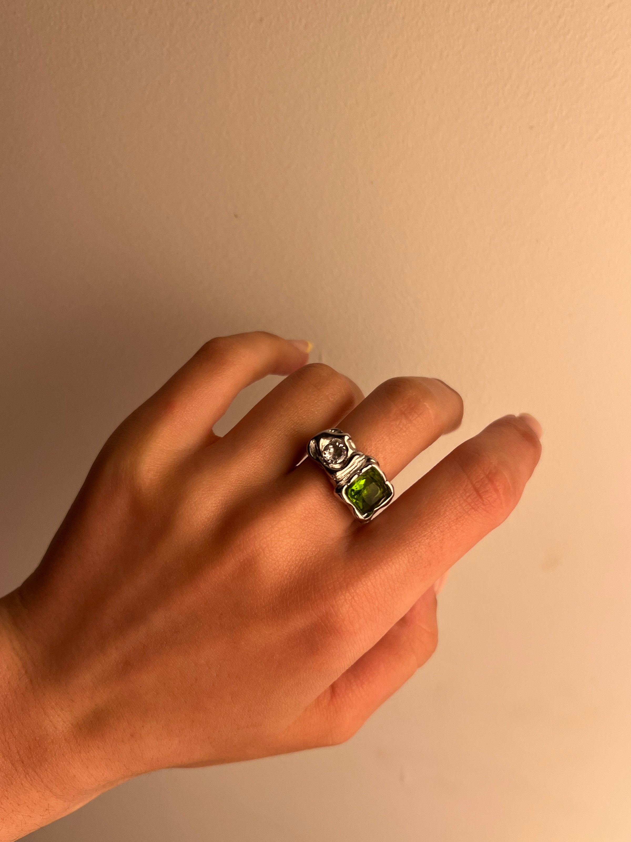 A hand wearing a silver ring with a large green gemstone, positioned against a neutral background.