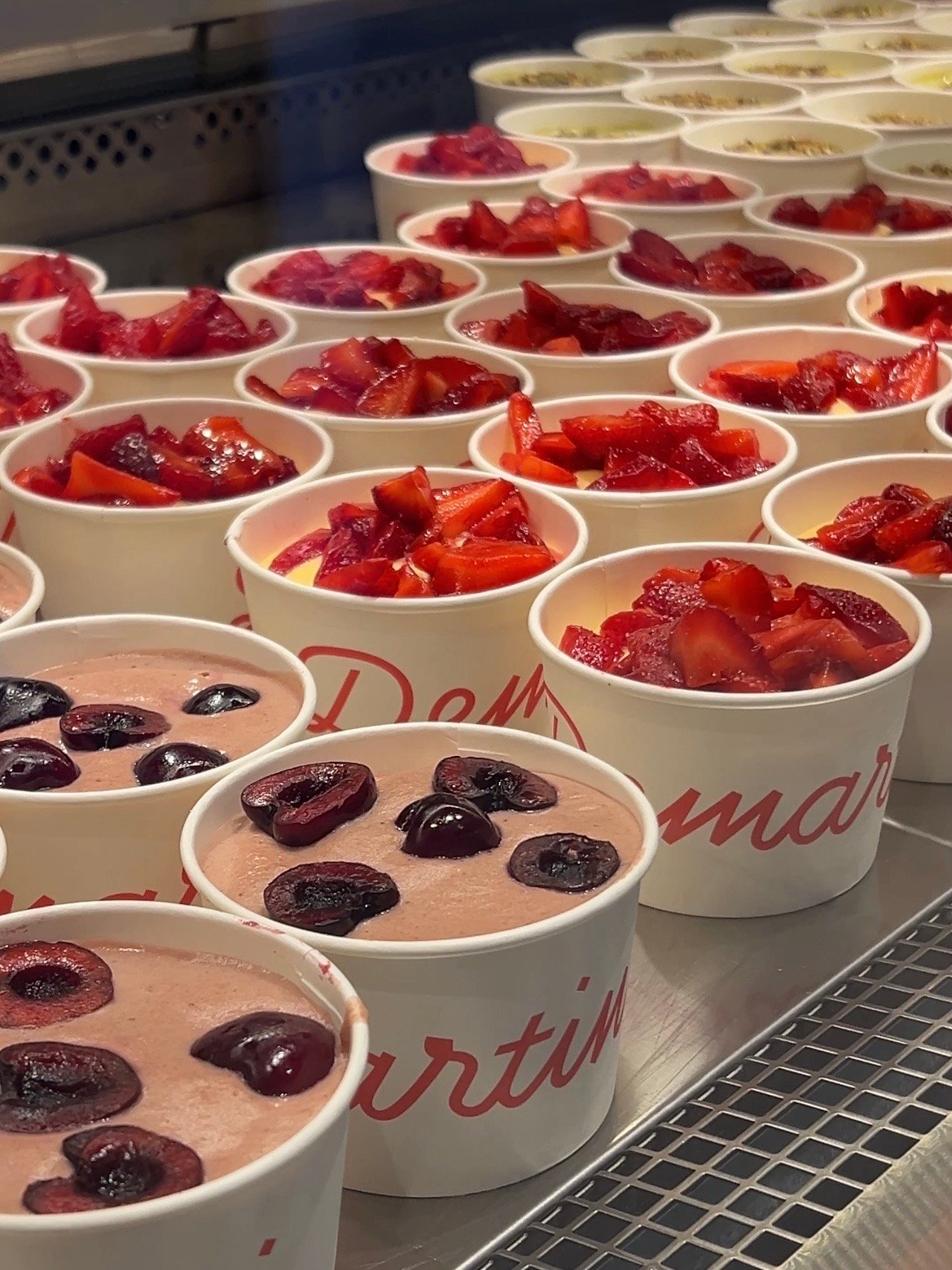 A display of dessert cups filled with various fruit toppings, including strawberries and cherries, arranged neatly in a glass case.