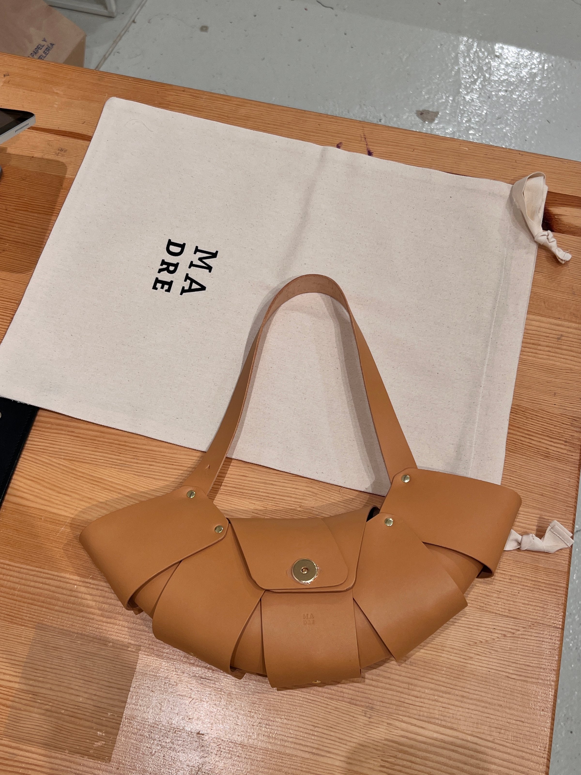 A stylish tan handbag with a unique folded design, placed on a wooden surface next to a canvas bag featuring the word 'MADRE'.