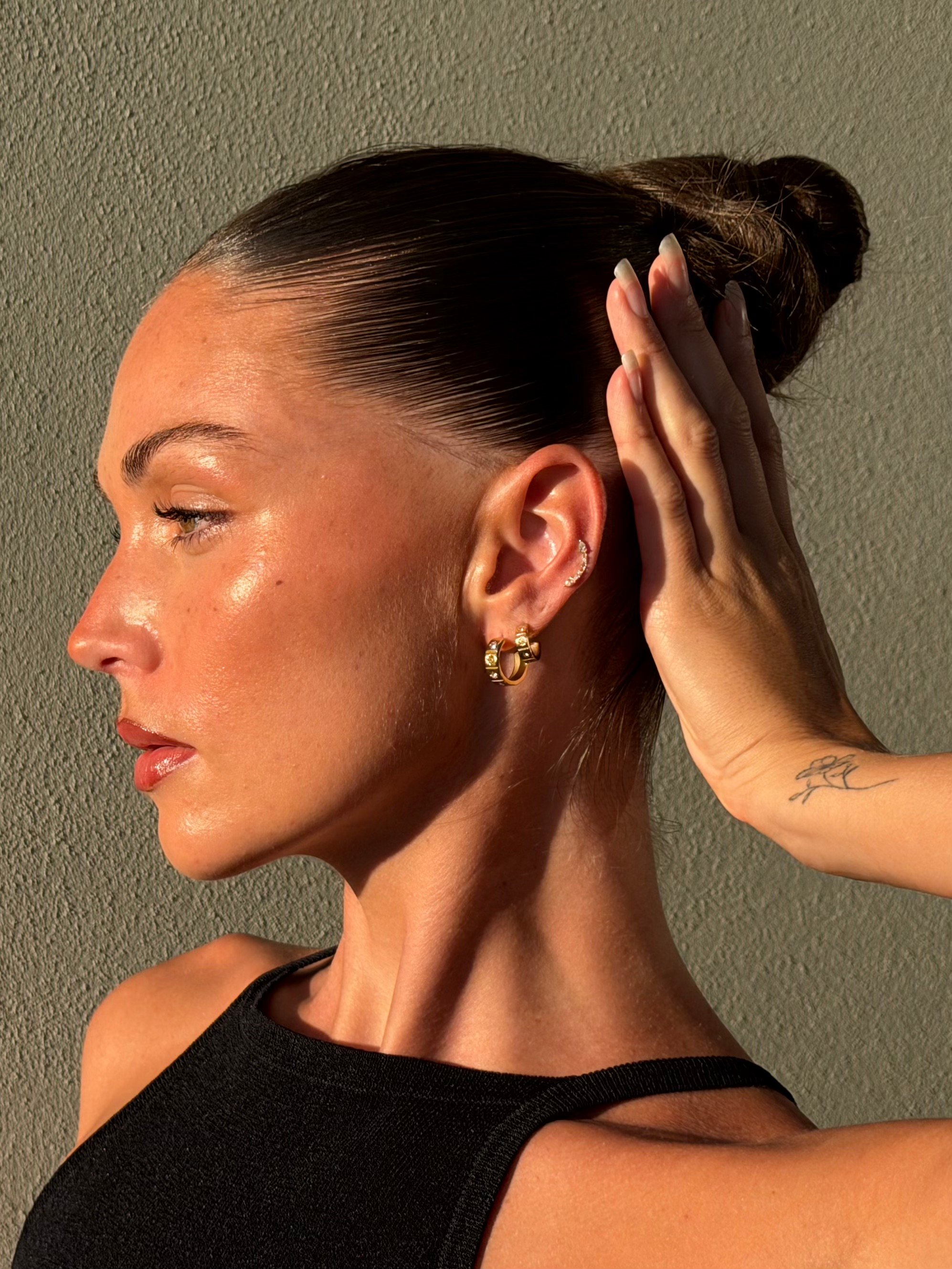 A profile portrait of a woman with slicked-back hair, showcasing her earrings and glowing skin against a textured background.