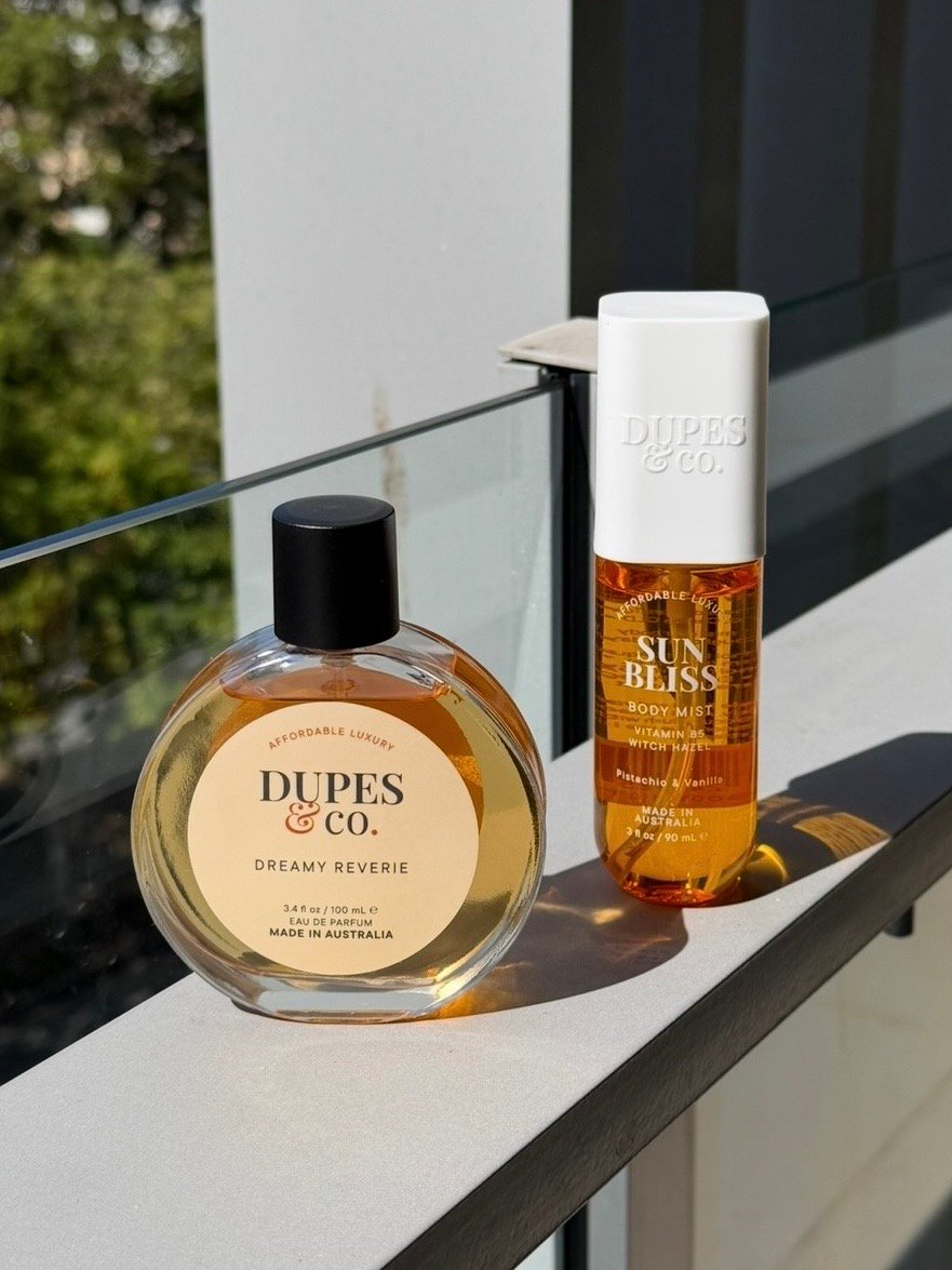 Two bottles of Dupes & Co. products displayed on a balcony railing, with greenery in the background.
