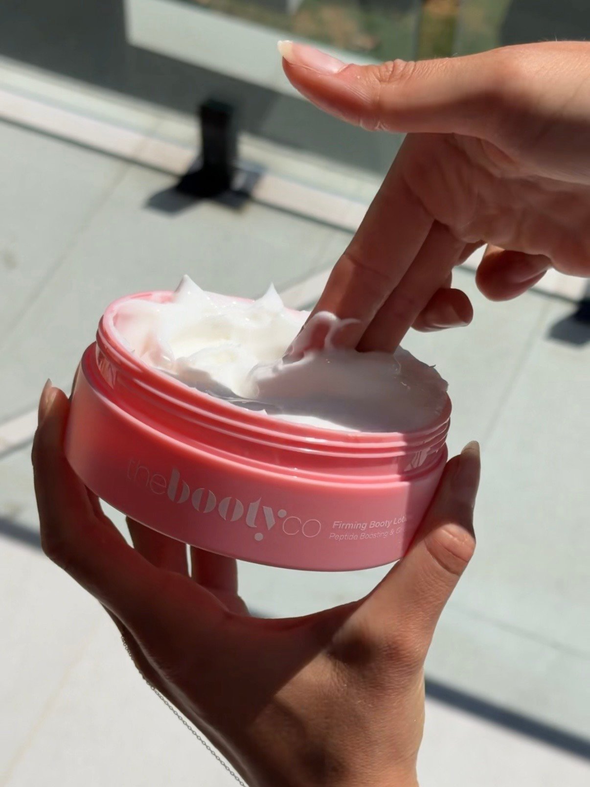 A hand applying cream from a pink container labeled 'the booty co' in a bright, sunlit setting.
