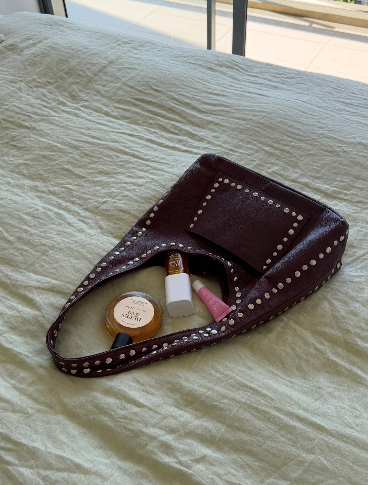 A maroon handbag with decorative studs, resting on a light-colored bedspread, surrounded by various beauty products.