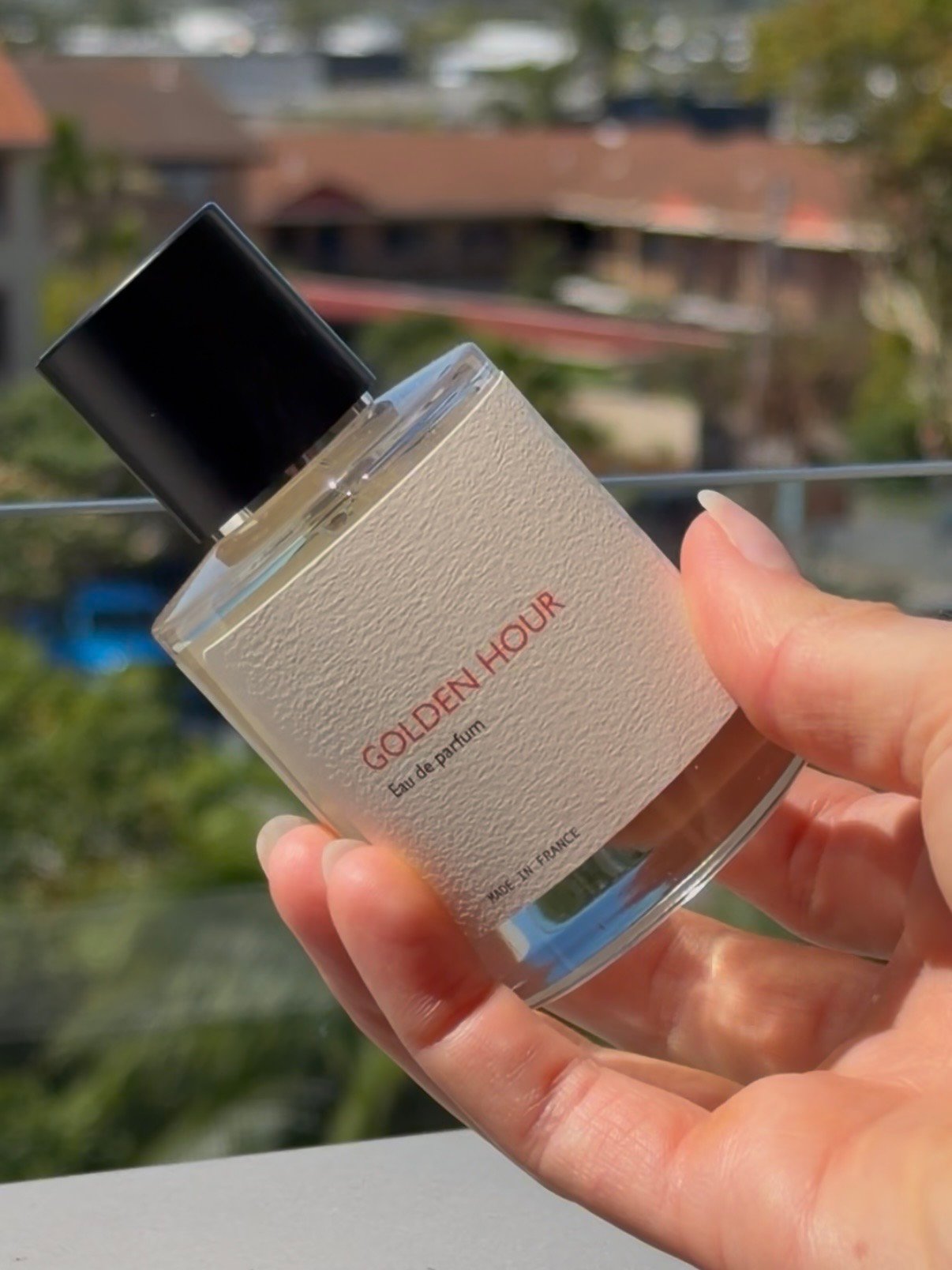 A hand holding a bottle of perfume labeled 'Golden Hour' against a blurred outdoor background.
